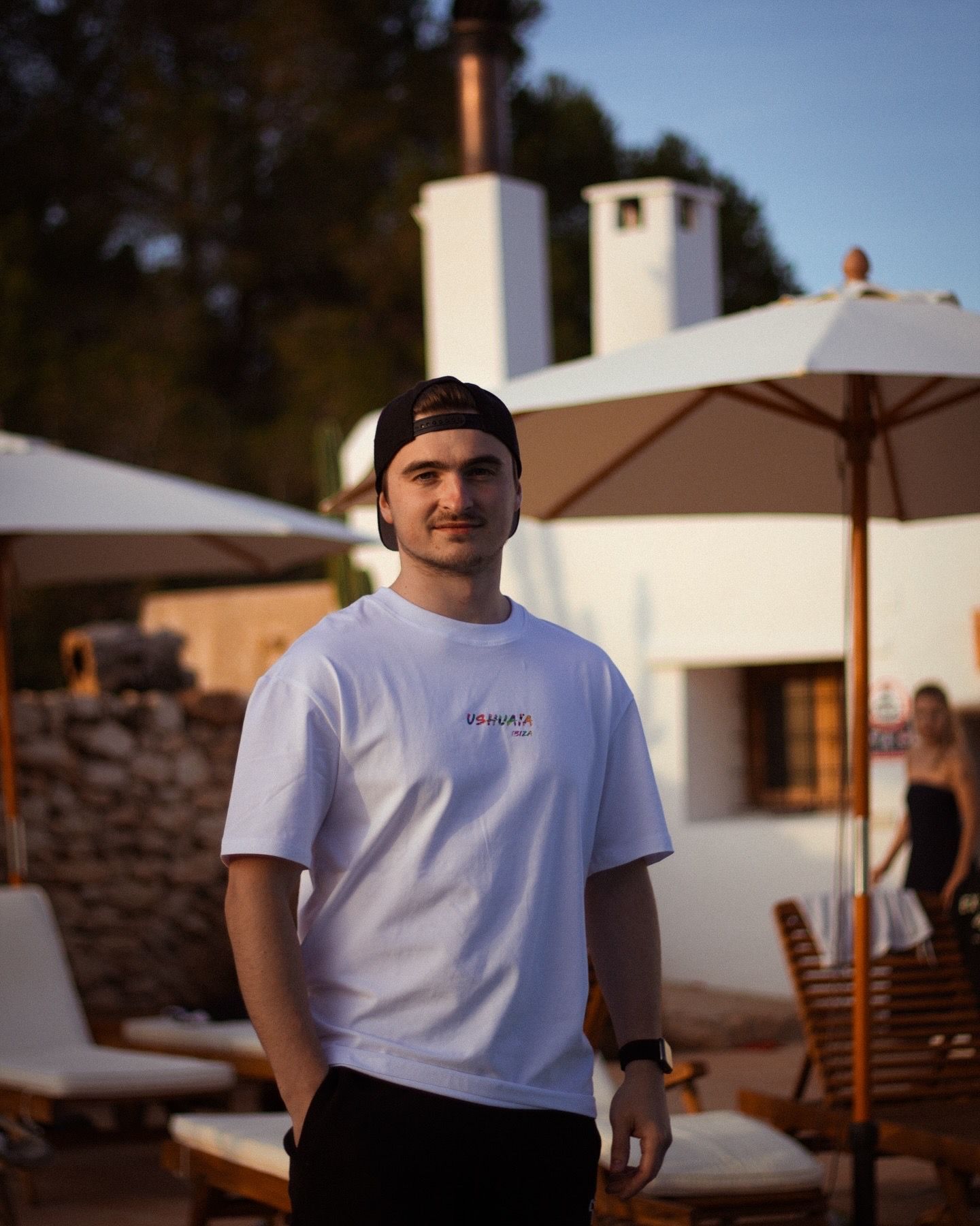 Person wearing white t-shirt and baseball cap standing outdoors near white building at sunset.