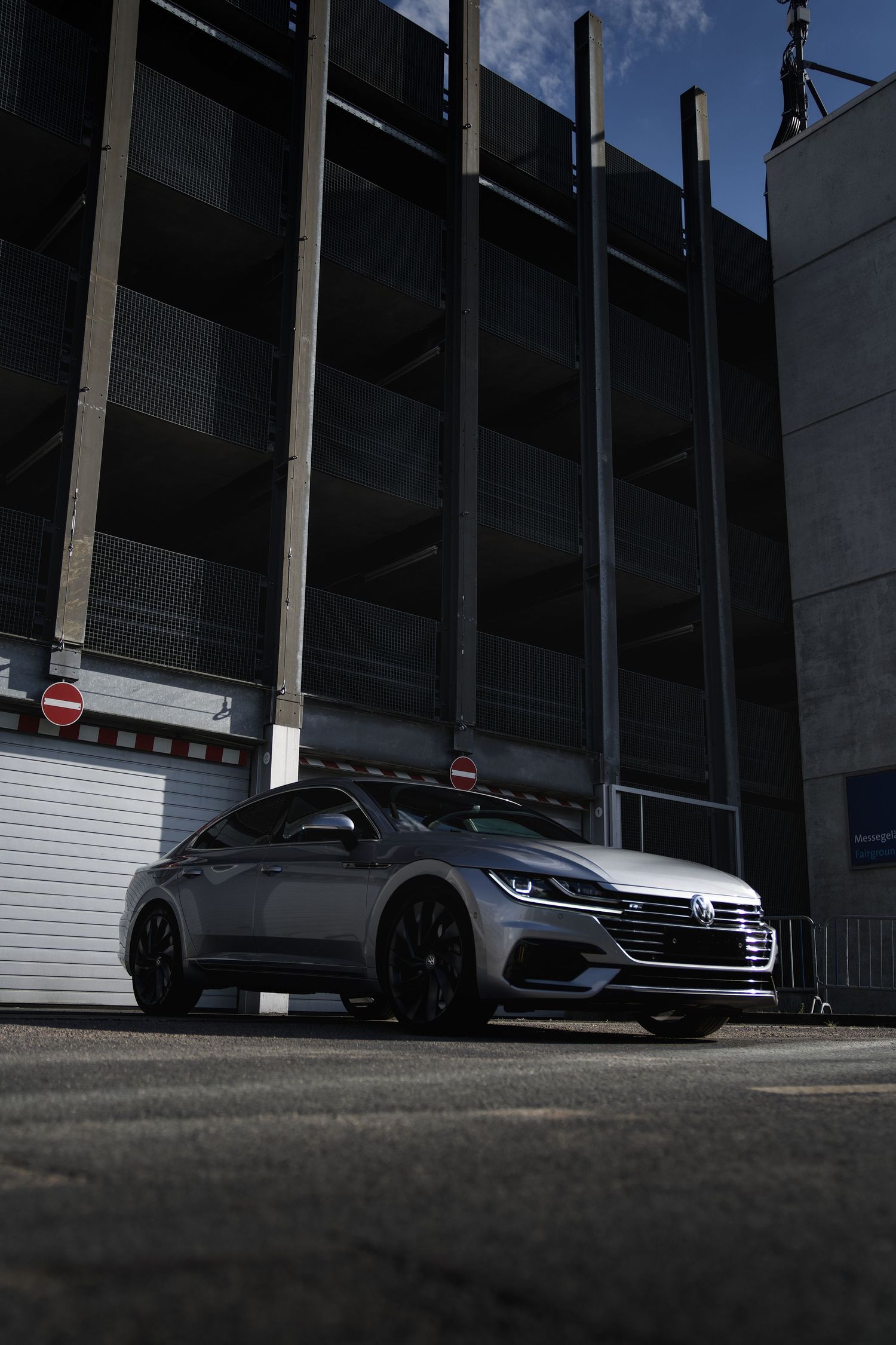 Silver Volkswagen Arteon parked in front of a multi-story parking garage on a sunny day.