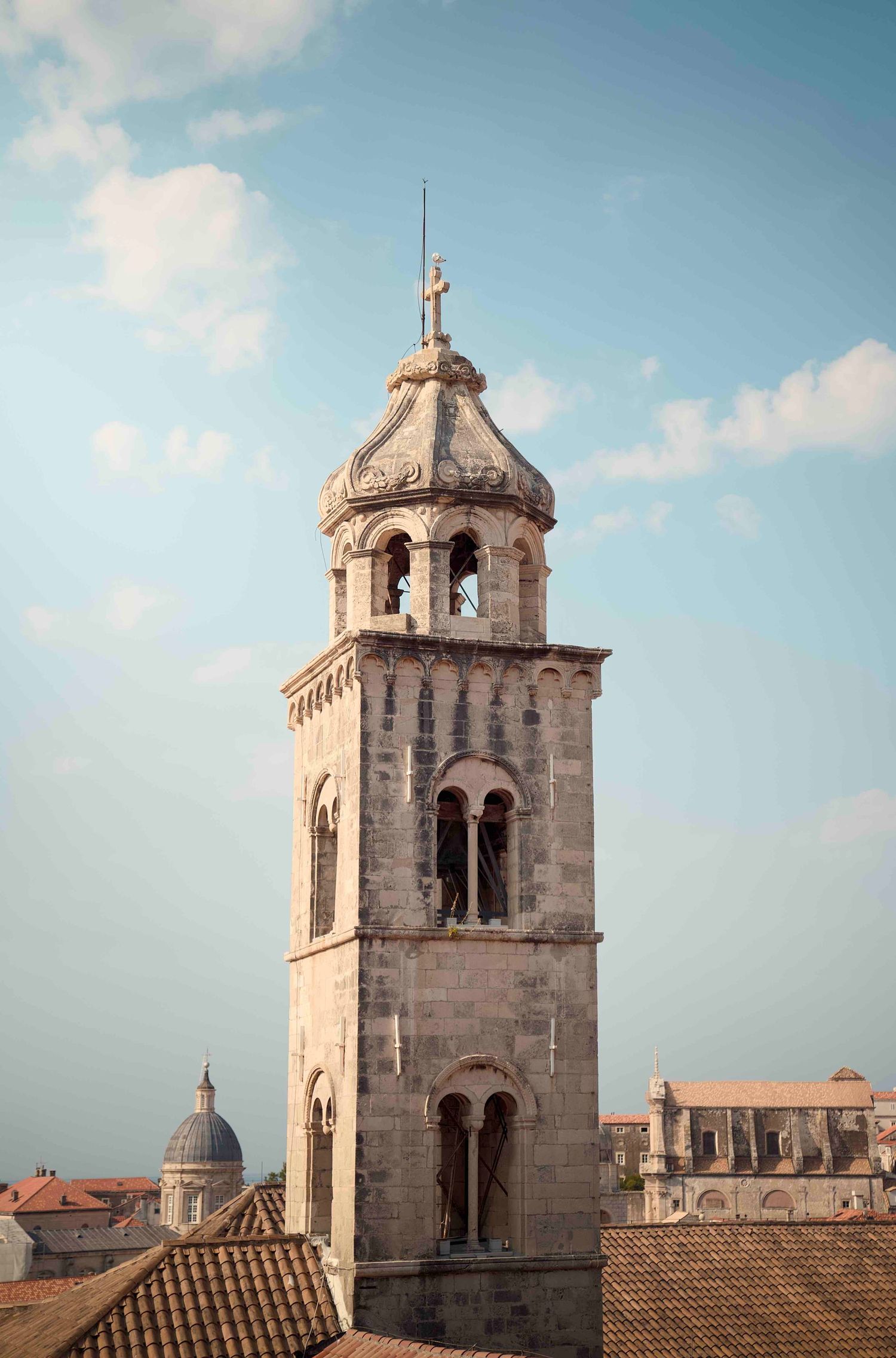 Historic stone bell tower with arched windows rises above terracotta rooftops against blue sky in Dubrovnik, Croatia.