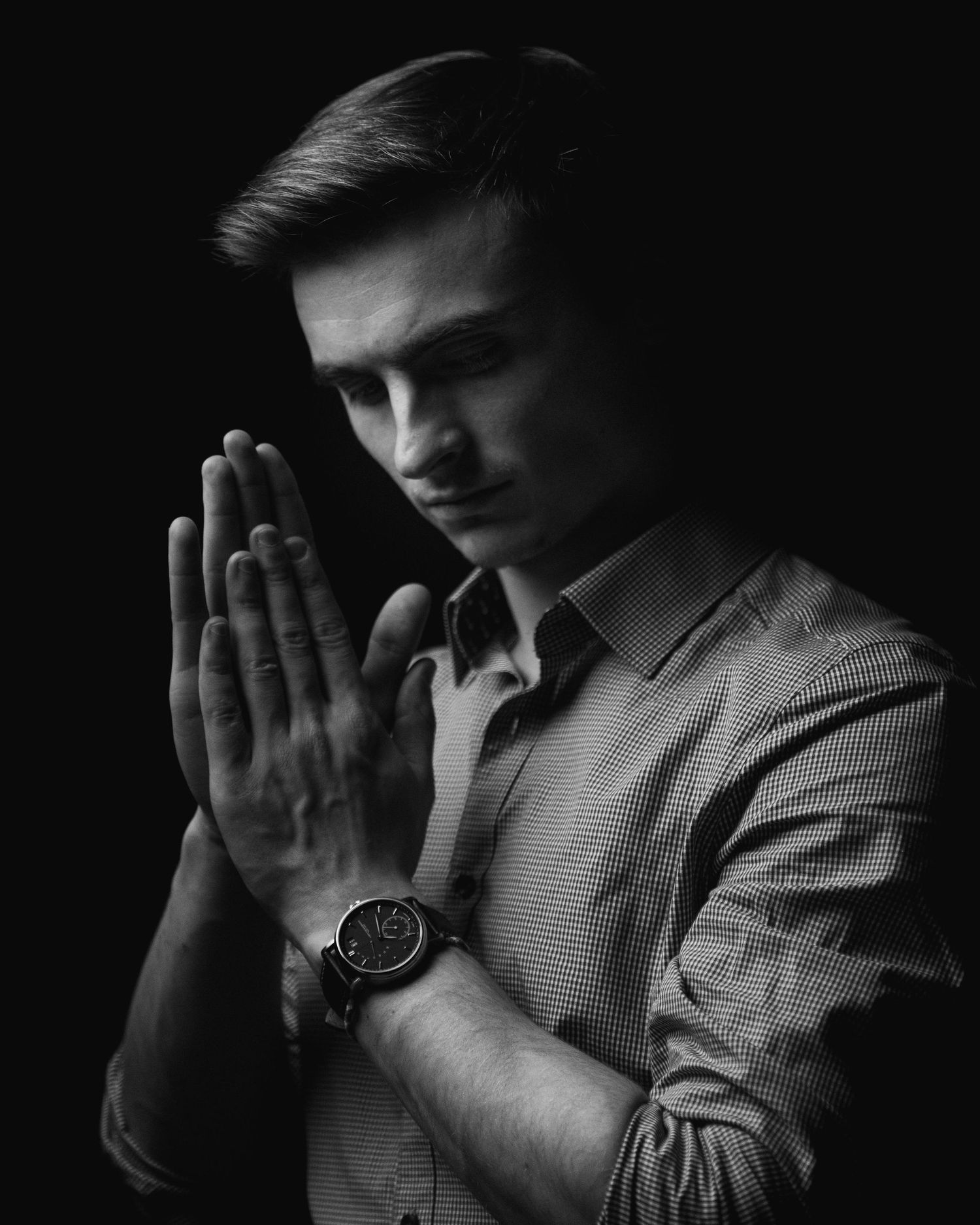 Dramatic black and white portrait of a person in prayer pose wearing a collared shirt and watch.