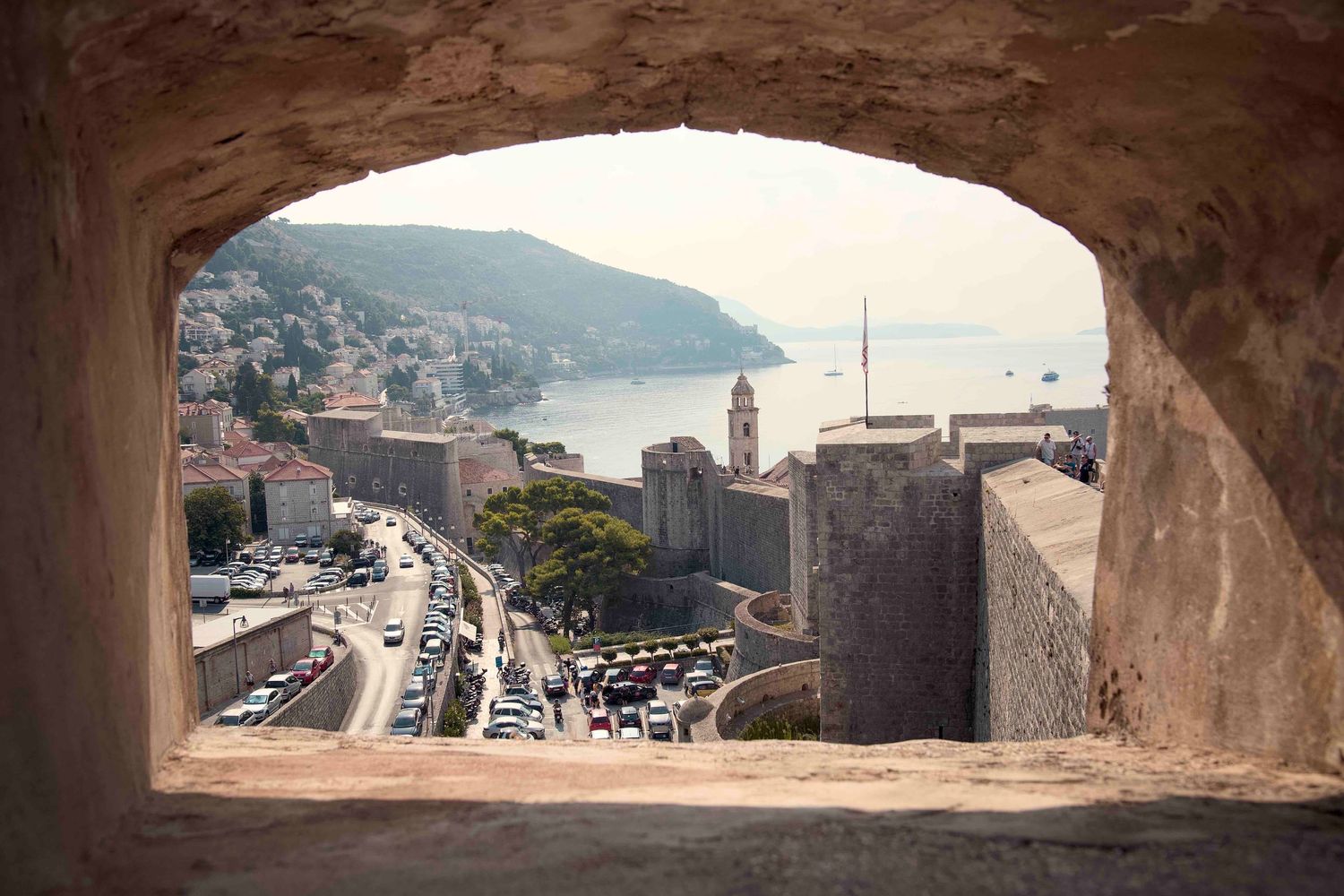 Panoramic views of a coastal Mediterranean city seen through stone archways of an ancient fortress wall.