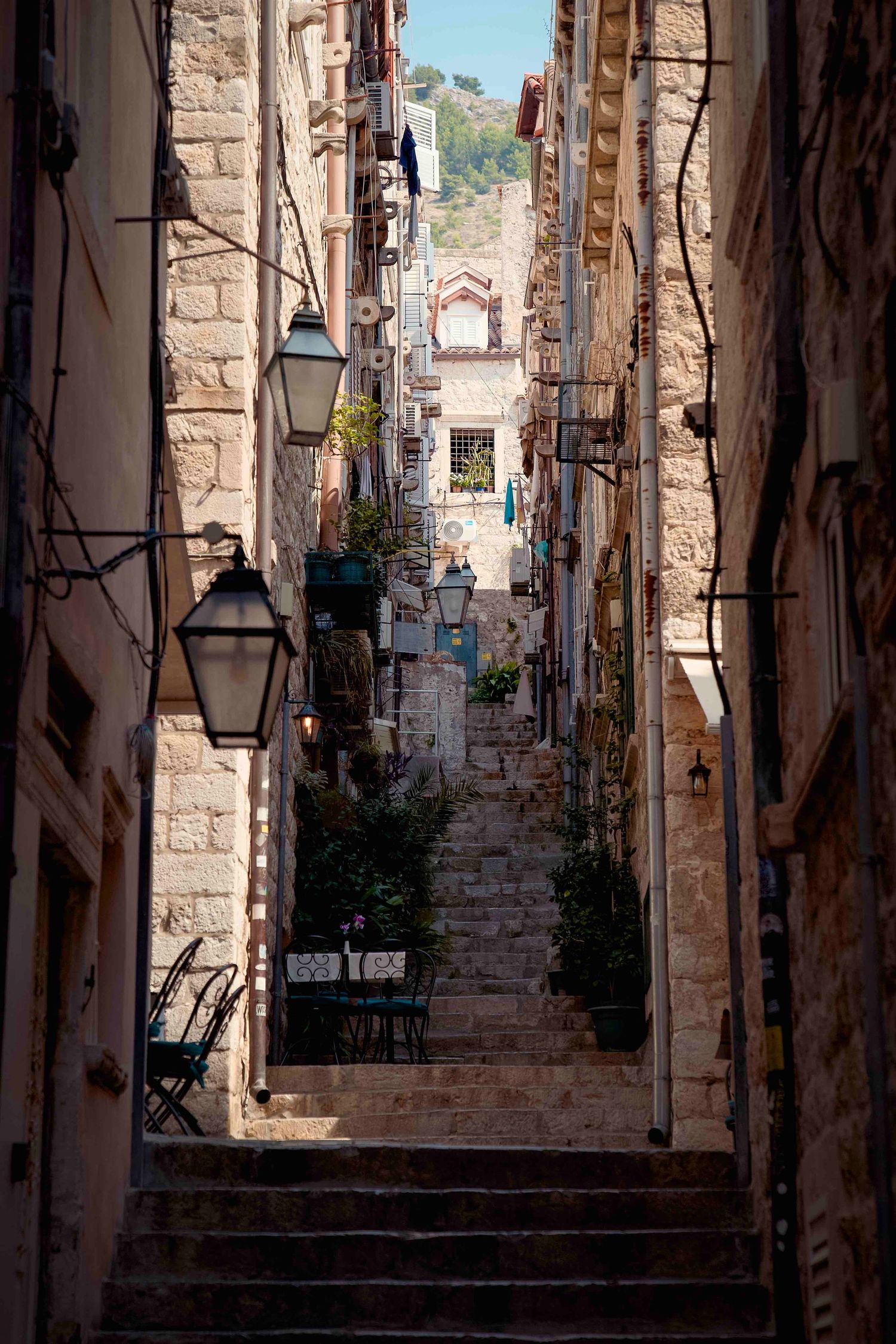 Narrow stone alleyway with steps in an old Mediterranean town featuring rustic architecture and hanging lamps.