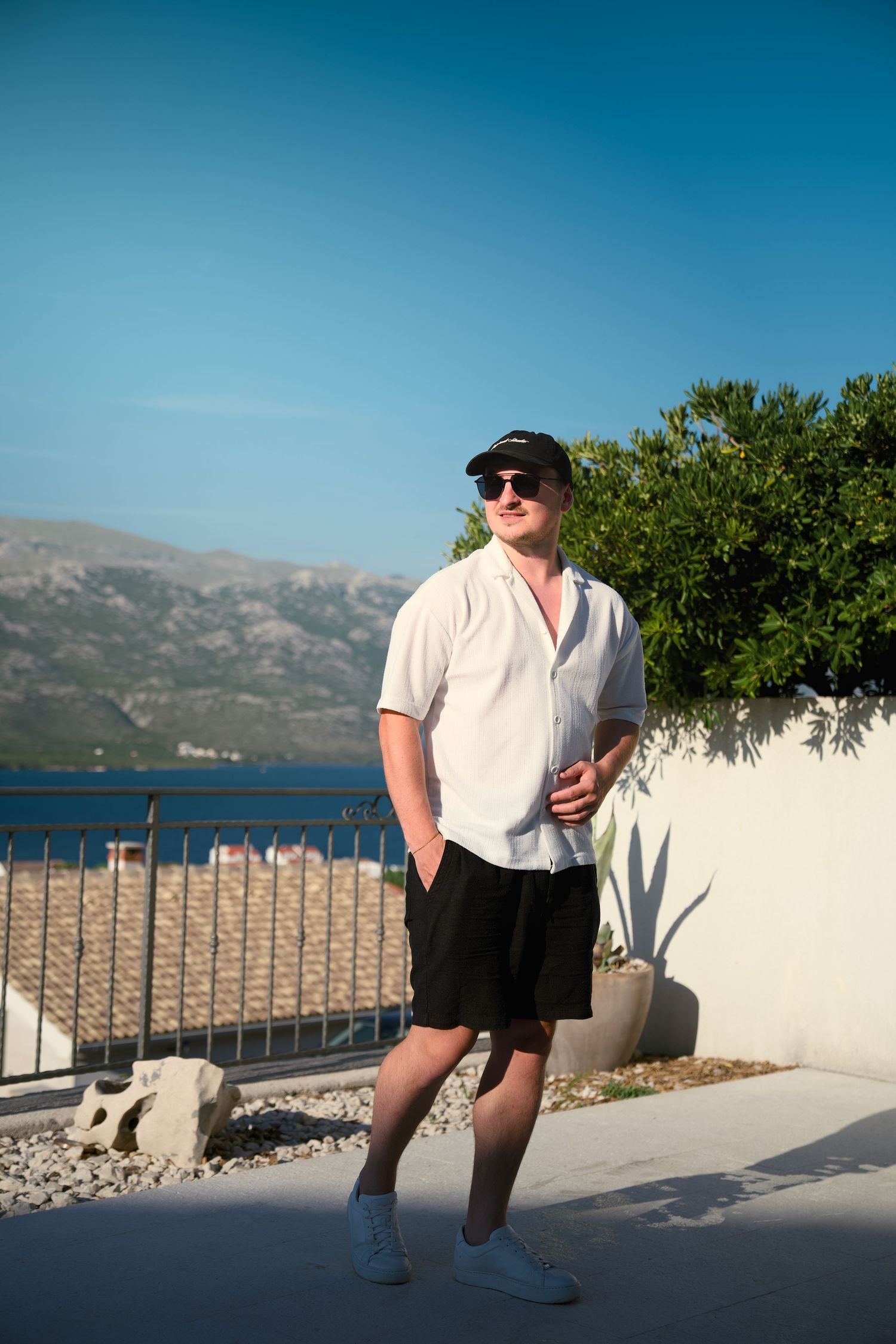 Man in white shirt, black shorts, cap and sunglasses standing on a terrace with sea and mountain views.