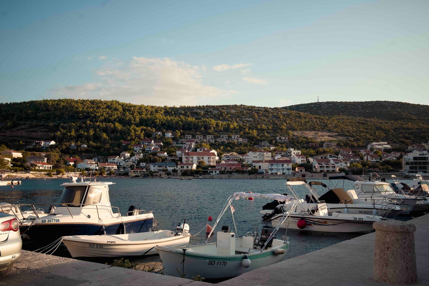 Boats docked in a Mediterranean harbor at sunset with hills and coastal village in the background.