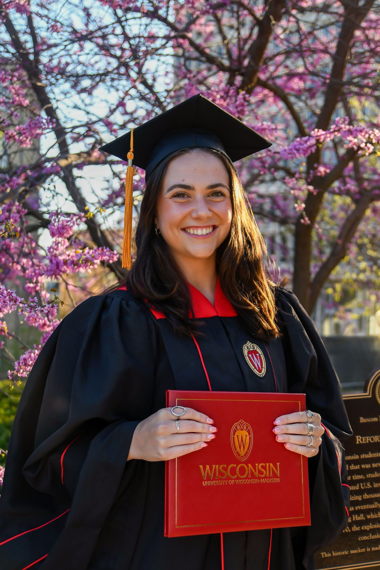 Student with cap and gown holding a diploma in front of a flowery tree.