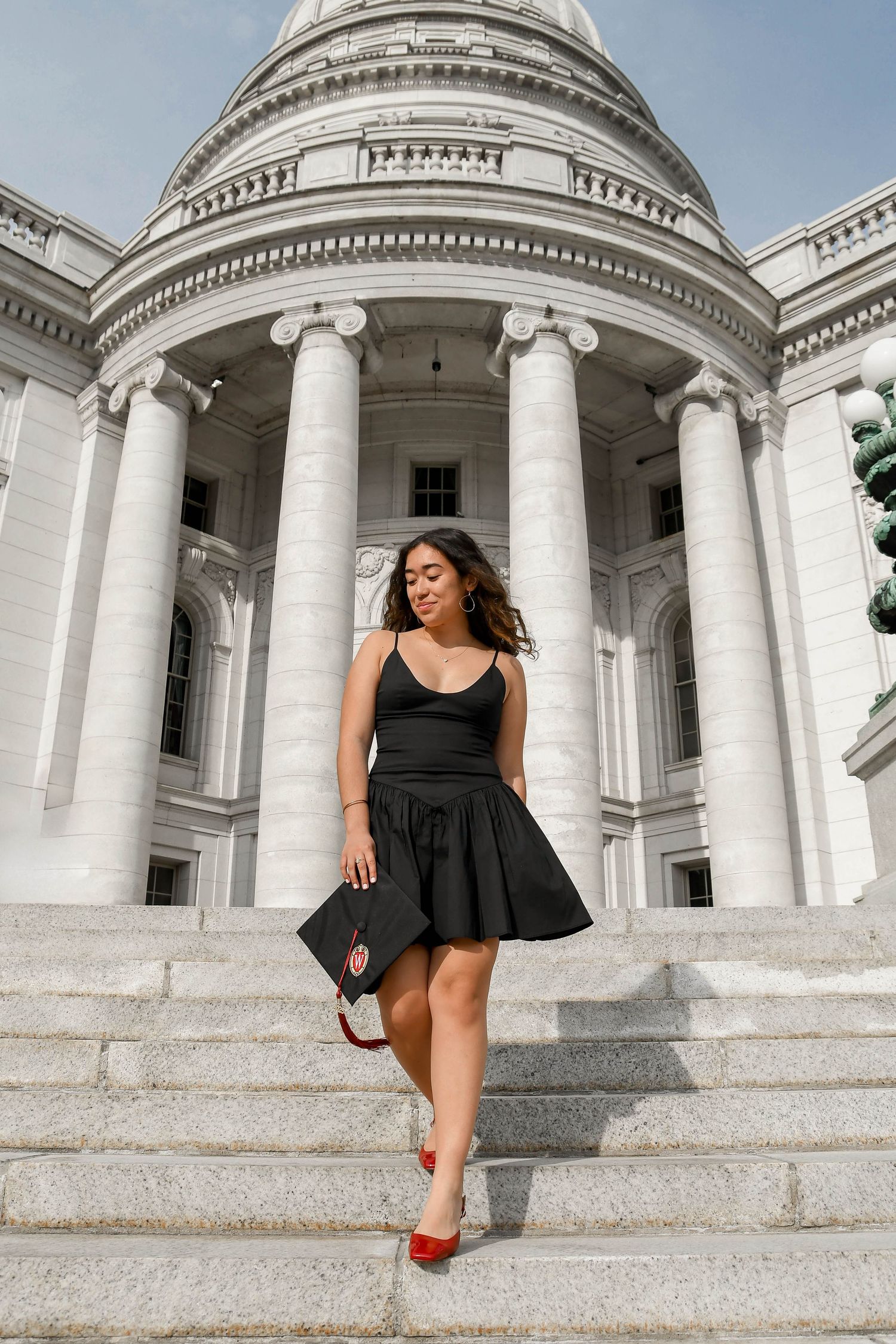 Girl walks down stairs of historical building holding graduation cap and looking away