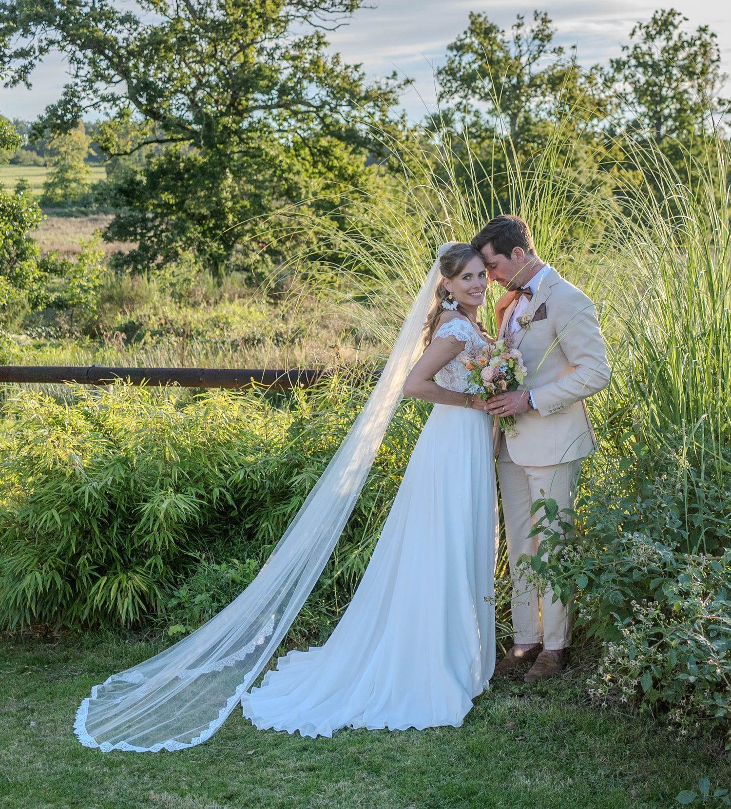 Bride and groom embracing in garden setting with flowing veil and natural greenery.