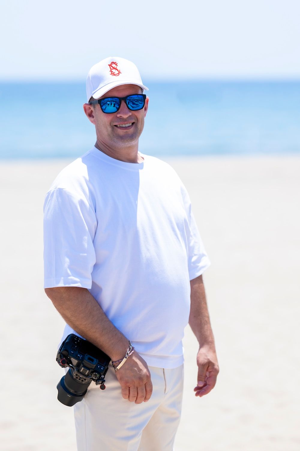 A person wearing a white shirt and baseball cap walks along a sunny beach on a bright summer day.