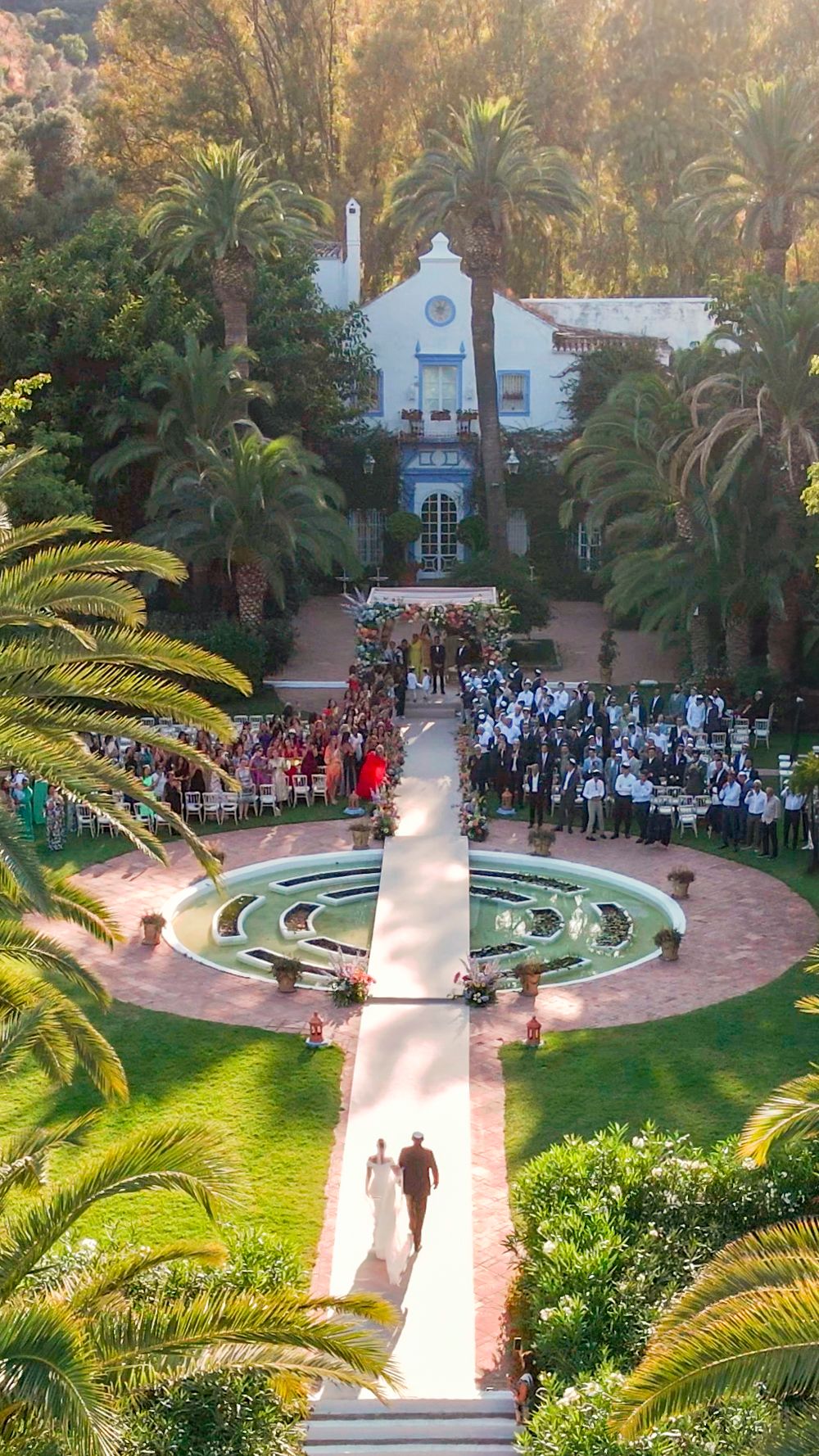 Aerial view of an outdoor wedding ceremony in a lush garden courtyard surrounded by palm trees and white architecture.