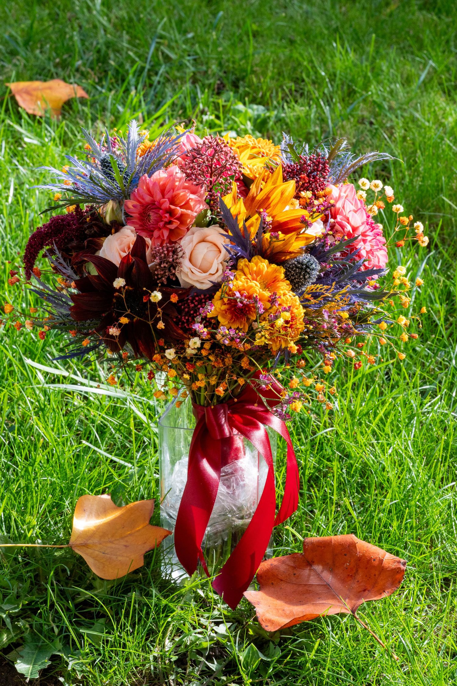 Vibrant autumn floral arrangement with red and orange blooms displayed on grass with fallen leaves in natural sunlight.
