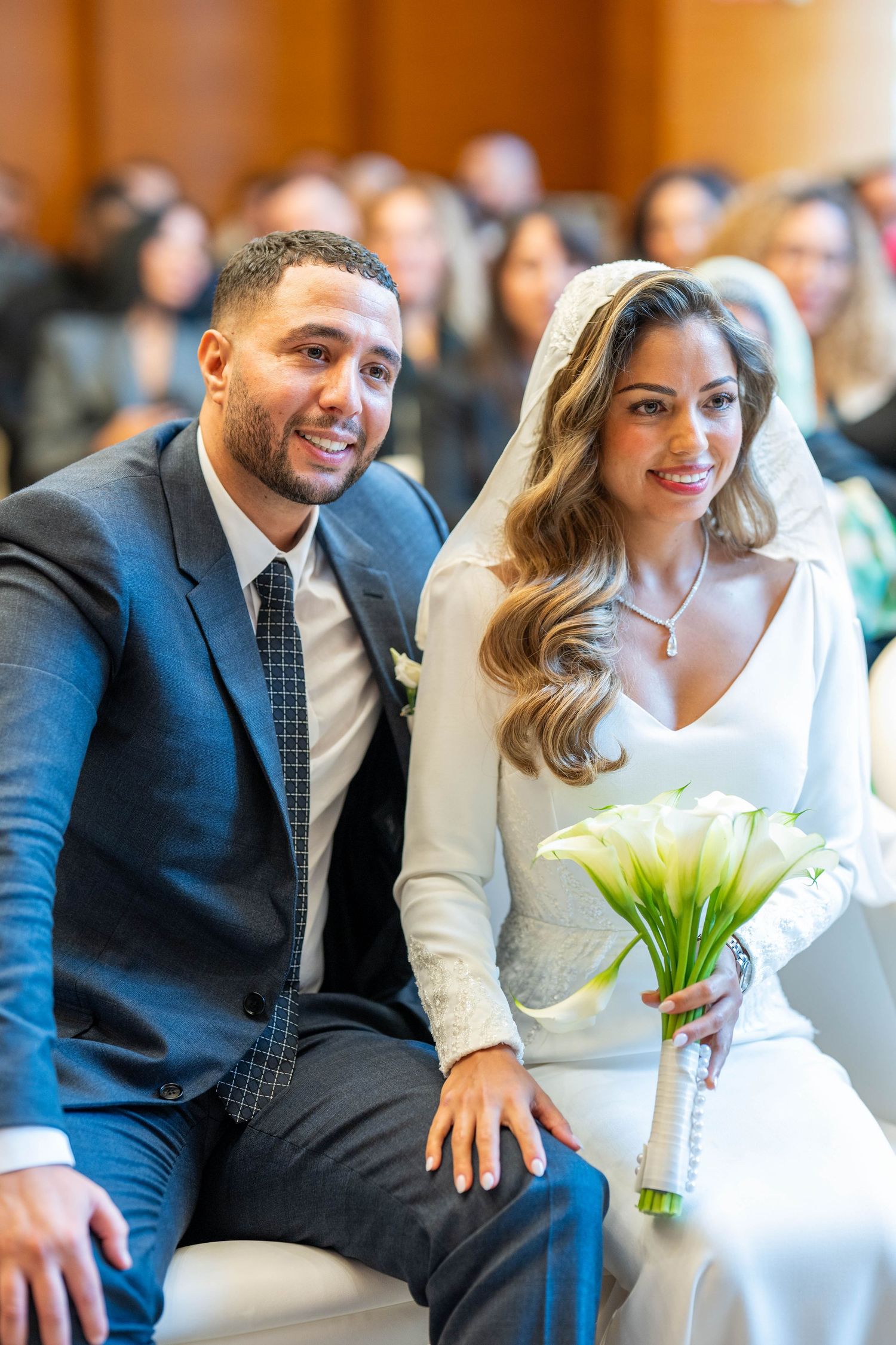 A couple in formal attire share a joyful moment together while seated, with a white floral bouquet visible.