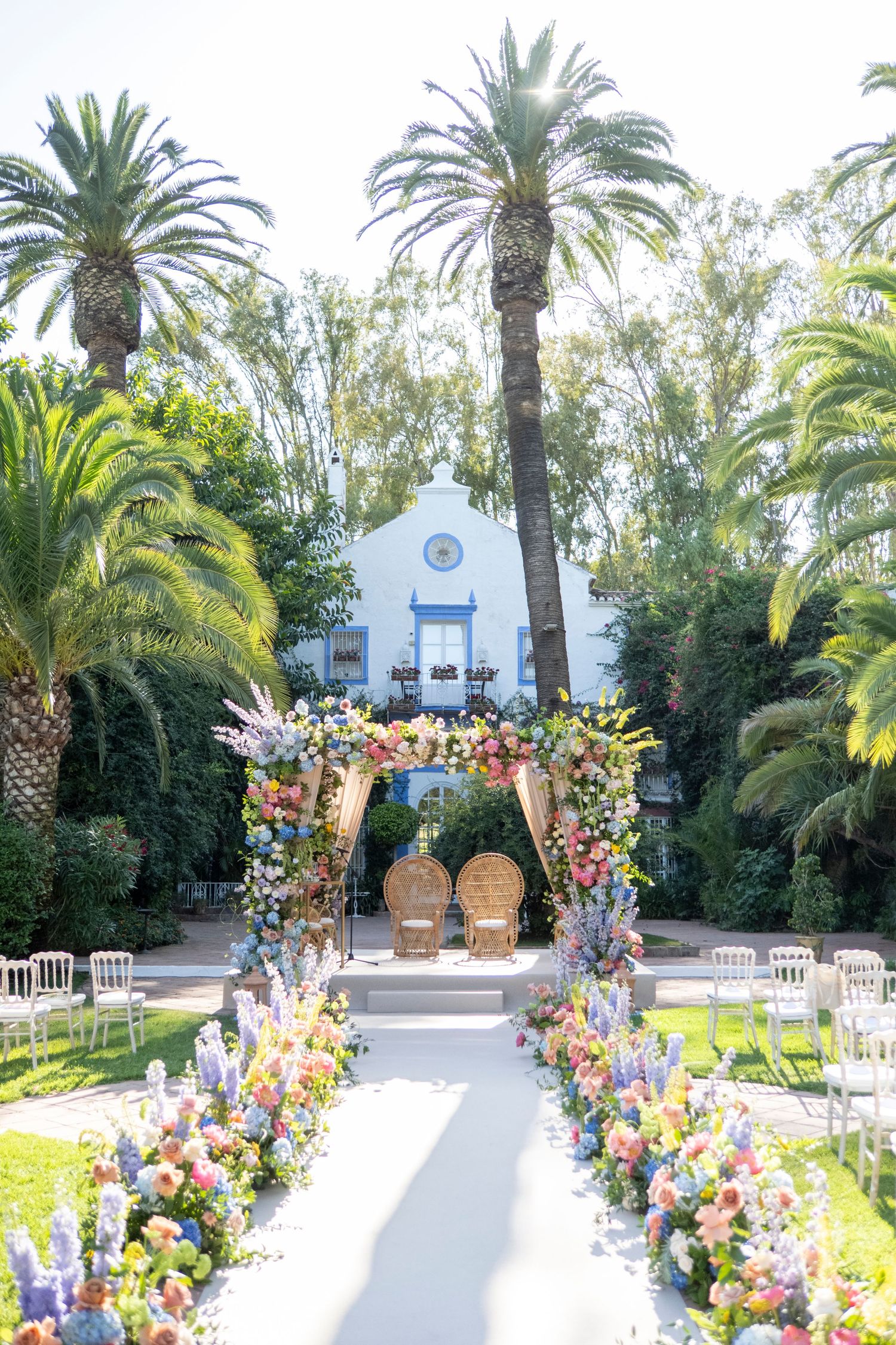 Outdoor wedding ceremony setup with floral arch, wine barrels and white aisle runner leading to a chapel among palm trees.