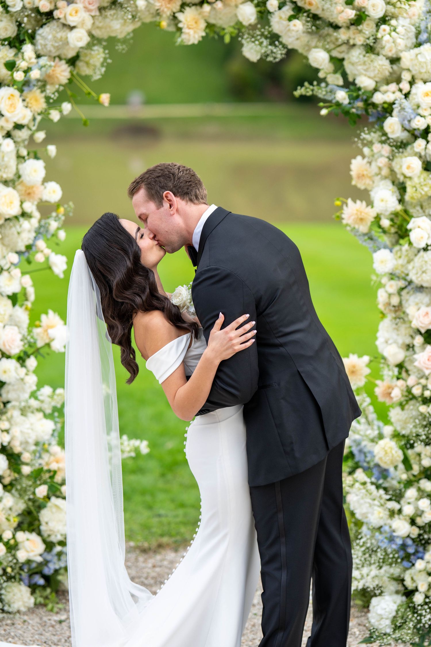 Bride and groom share a romantic kiss under a beautiful floral arch of white blossoms in an outdoor garden setting.