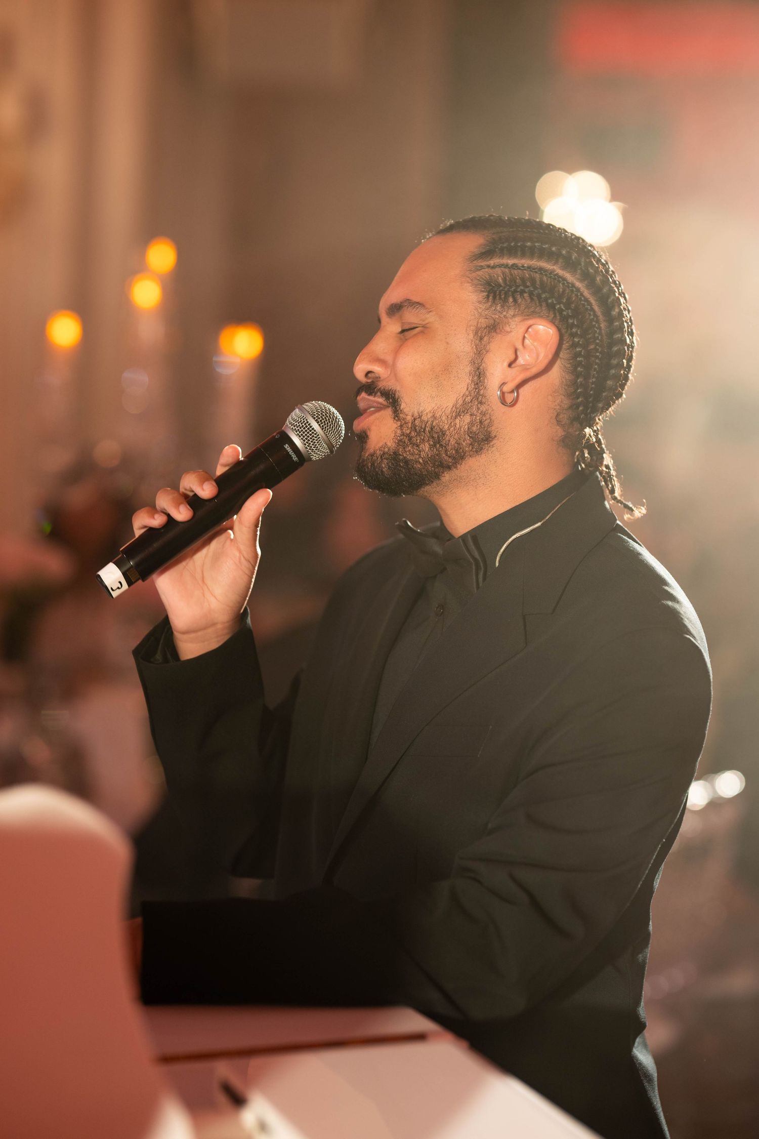 A performer in black attire sings passionately into a microphone during a live performance with warm lighting in the background.