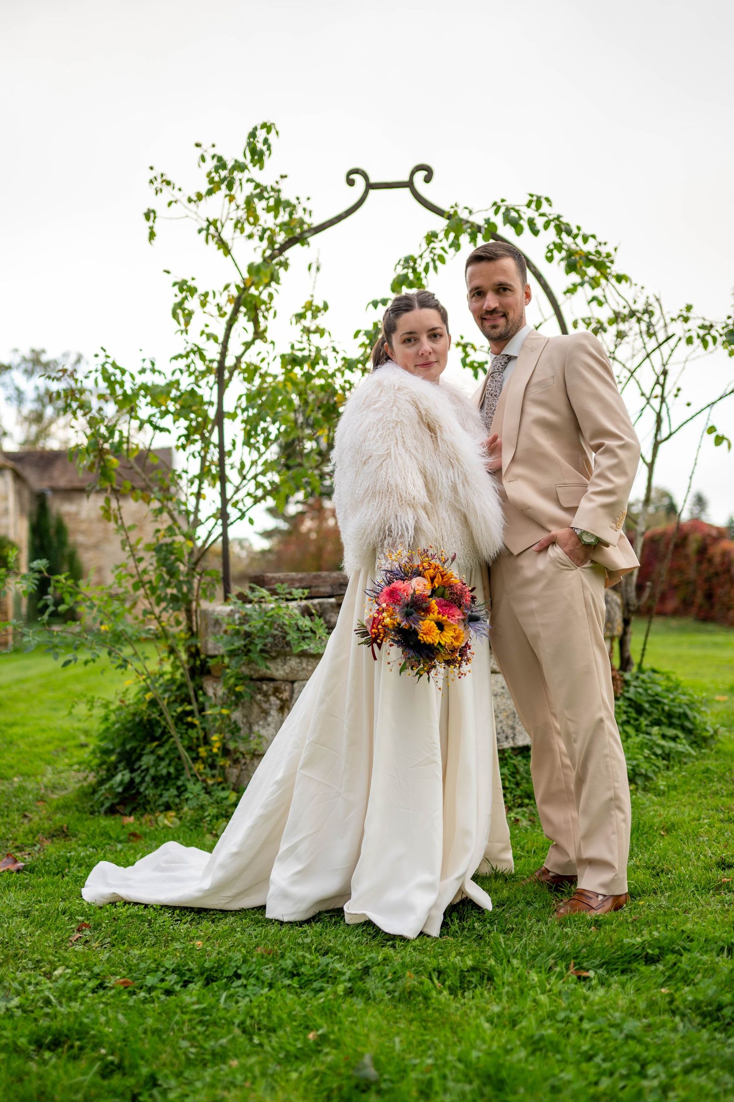 A wedding couple poses in a garden setting with the bride wearing a white dress and fur stole holding an autumn bouquet.