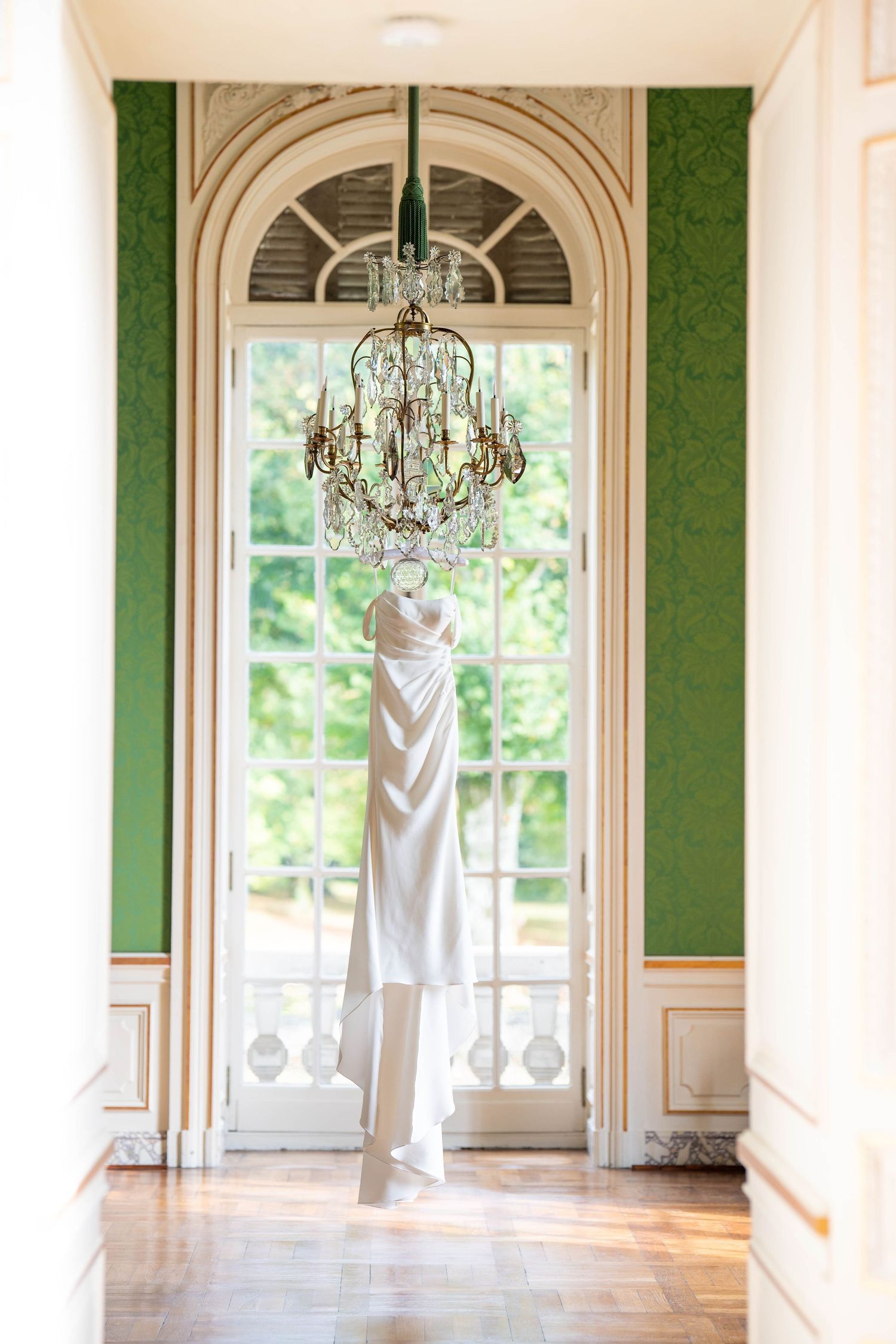 Elegant wedding dress hangs in a grand green room with crystal chandeliers and arched windows.