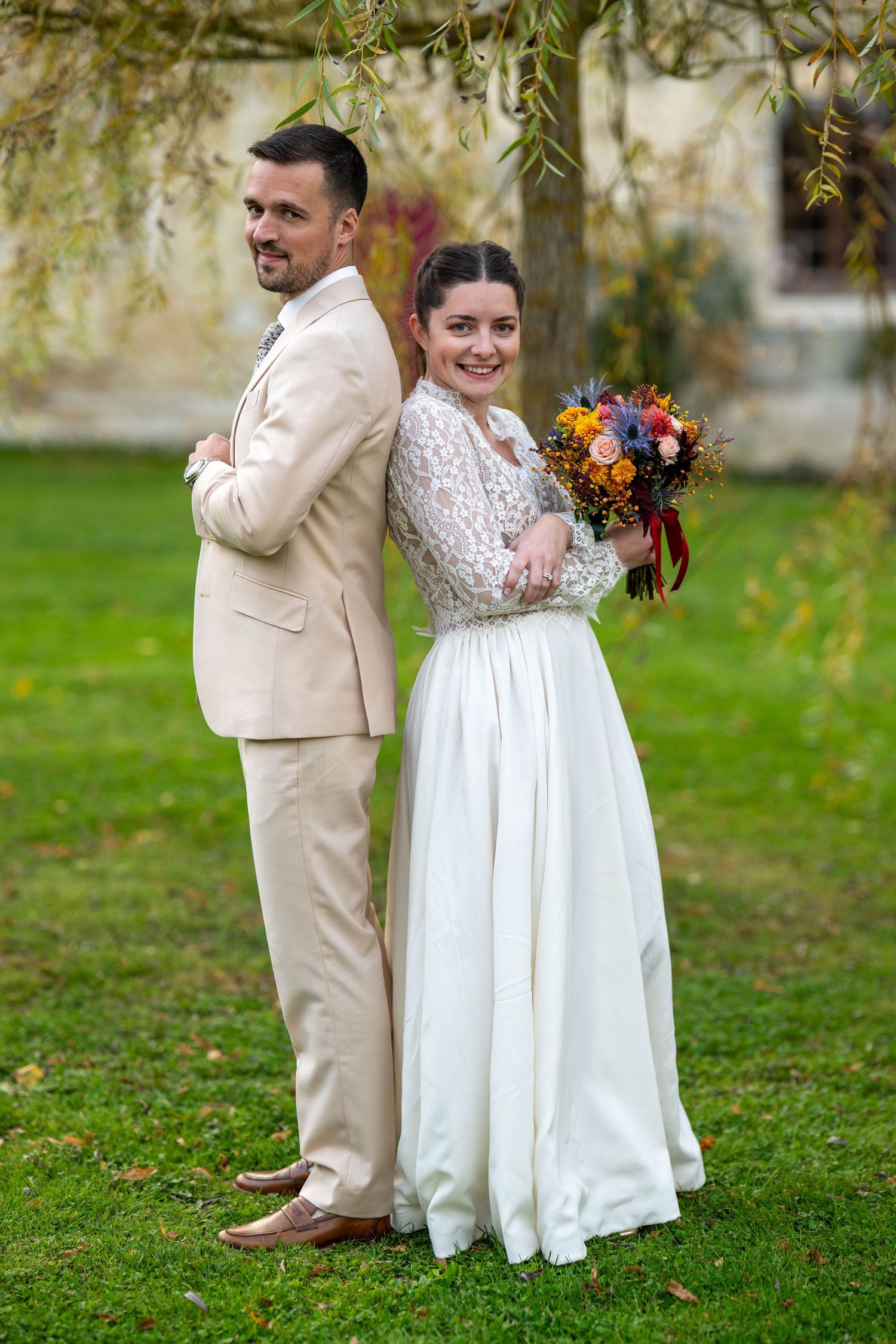 A couple poses in wedding attire with a fall bouquet in front of a garden setting.