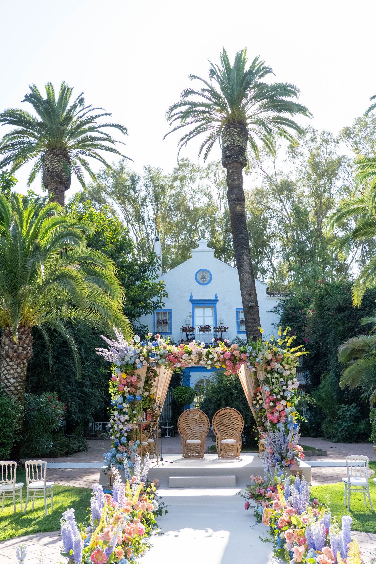 Outdoor wedding ceremony setup with floral arch and palm trees in front of a white church building.