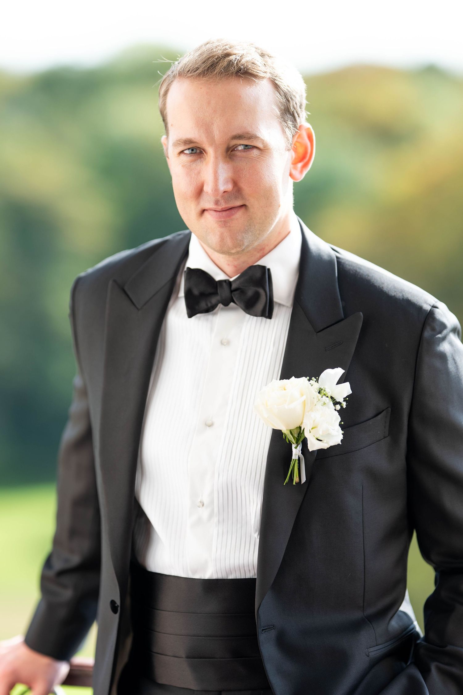 A groom in a classic black tuxedo and white bow tie poses outdoors on his wedding day.