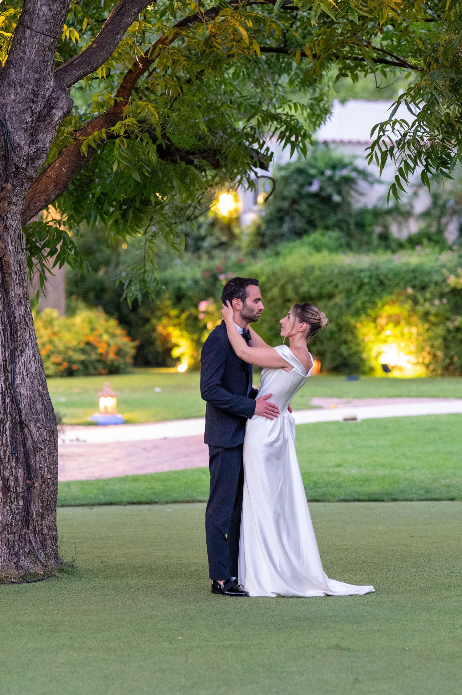 A couple shares a romantic moment dancing together on a lush green lawn at sunset with trees in the background.