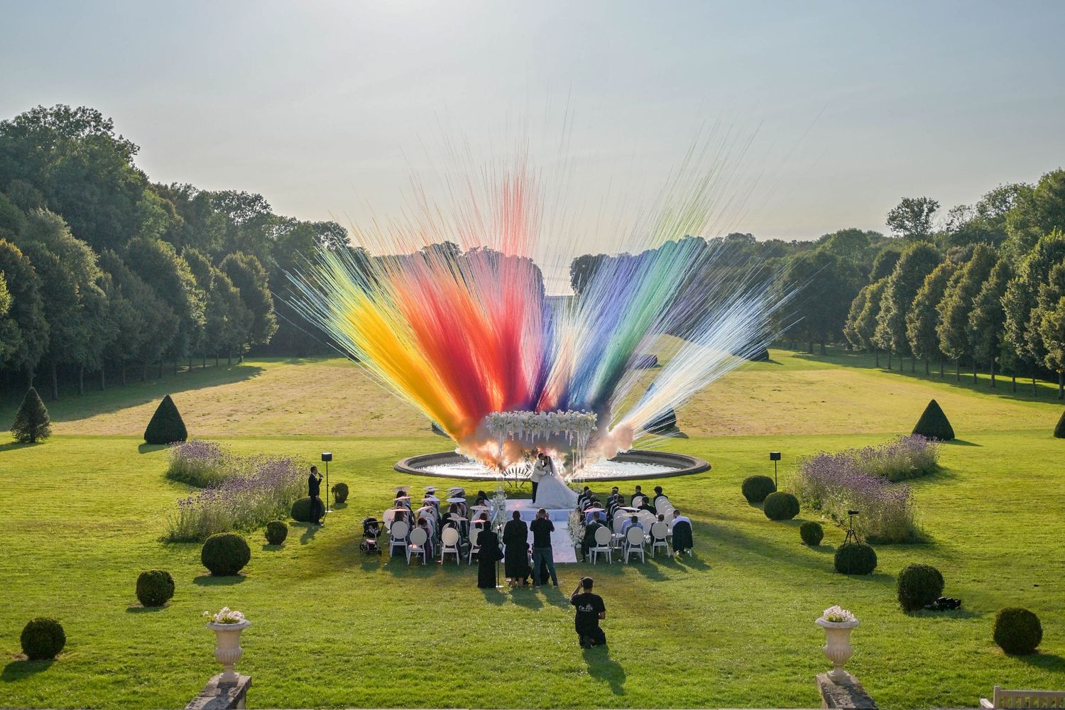 Colorful rainbow smoke fills the sky over a green field during an outdoor celebration.