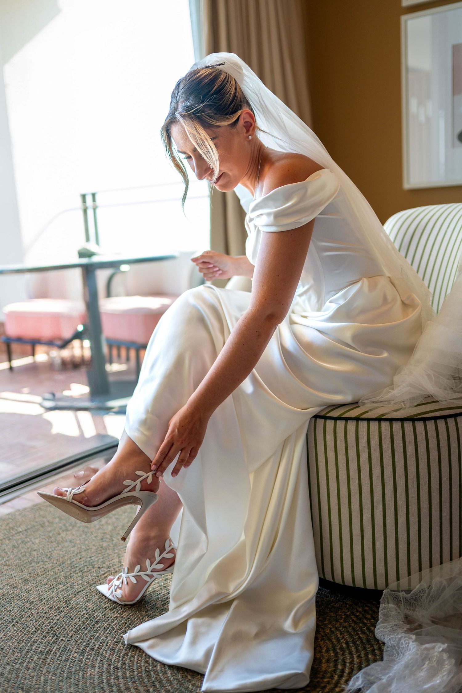 A bride in an elegant white wedding dress sits on a striped couch while putting on sparkly heels.
