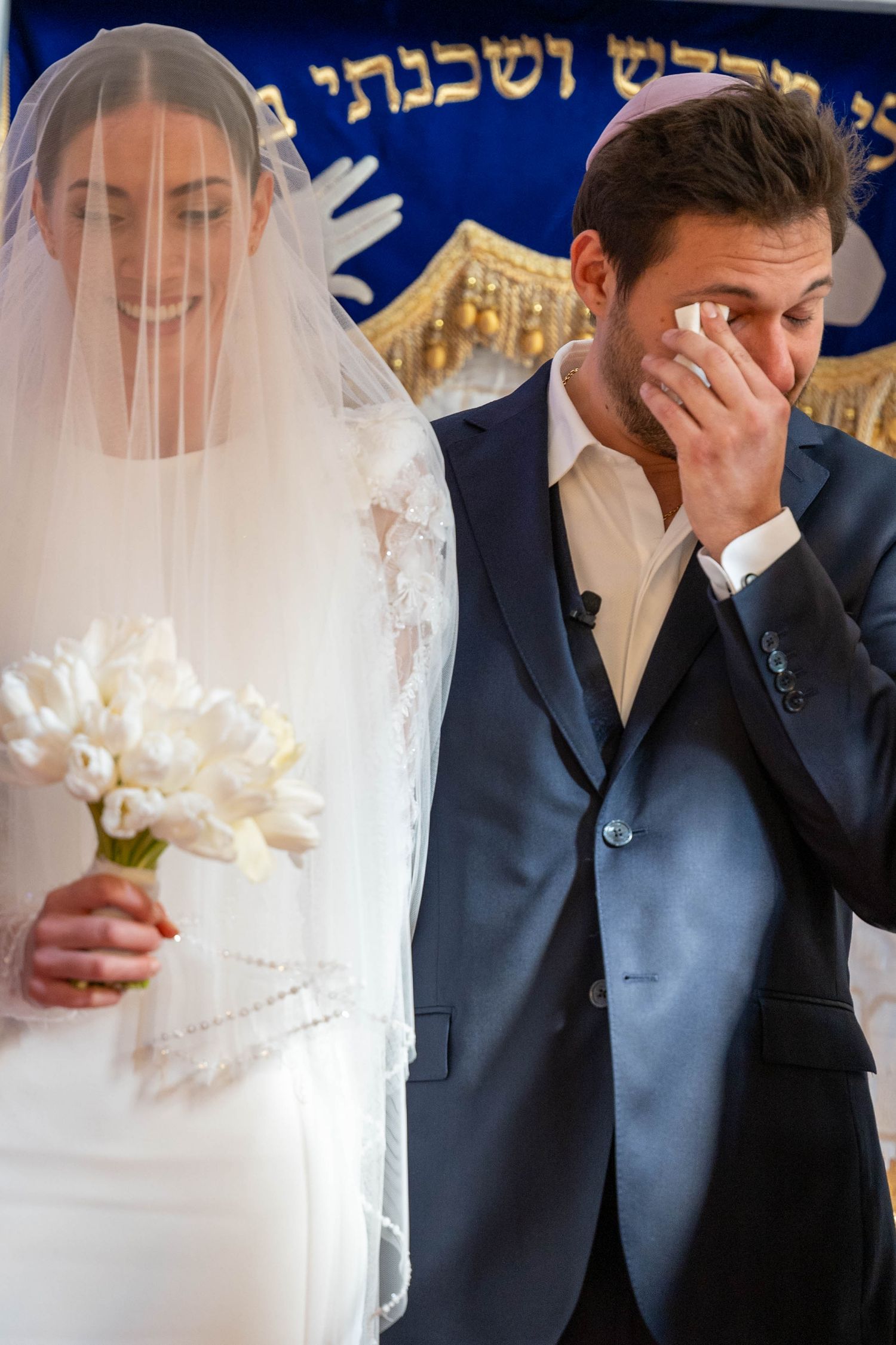 Emotional wedding ceremony moment under traditional Jewish chuppah with white floral decorations.