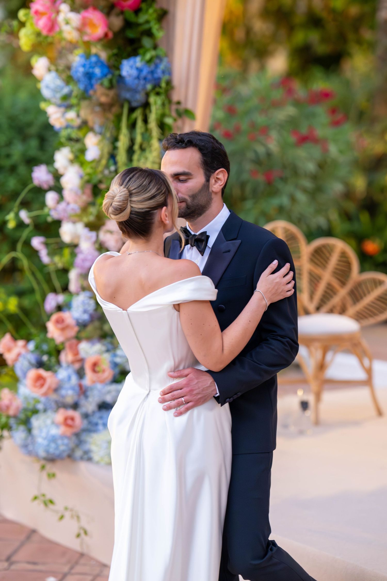 A couple shares a tender embrace during a wedding celebration surrounded by colorful floral arrangements.