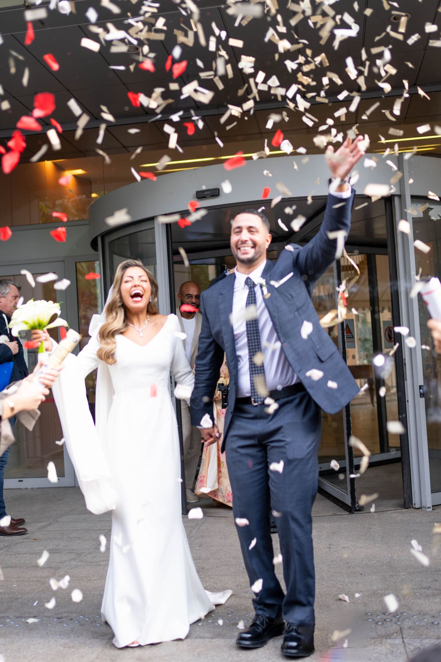 A wedding couple celebrates with confetti being thrown in the air outside a building entrance.