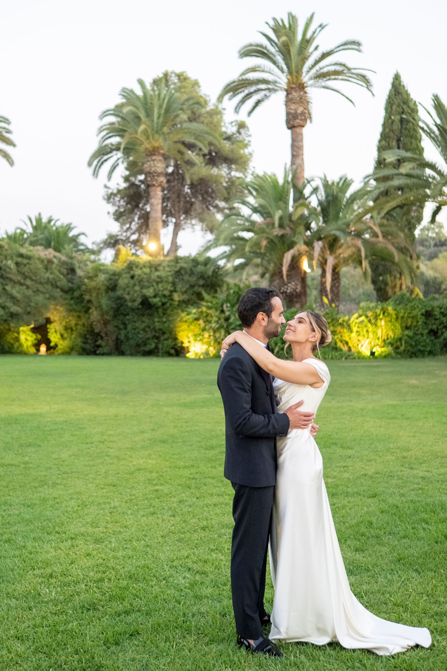 A couple in wedding attire dances together on a lush green lawn surrounded by palm trees at sunset.