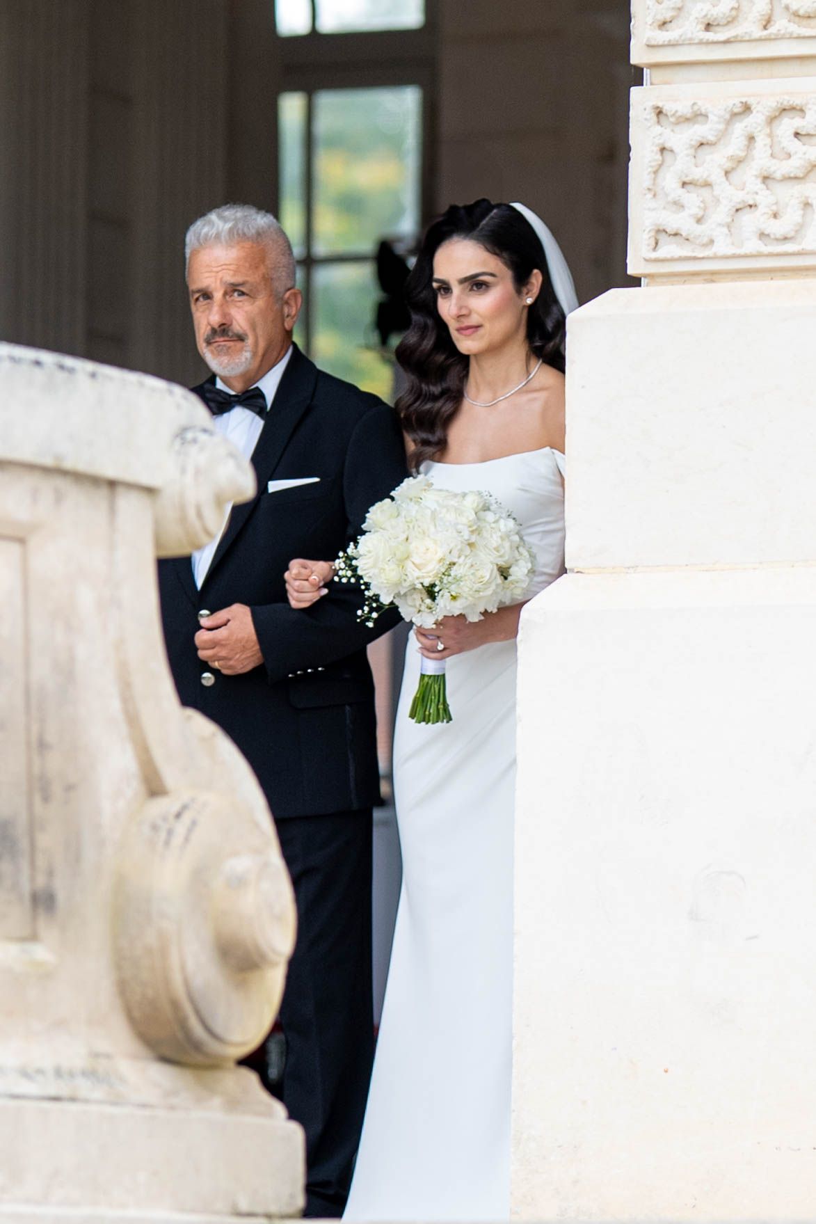 A bride in a white gown and groom in a black suit share an intimate moment against an ornate stone wall.
