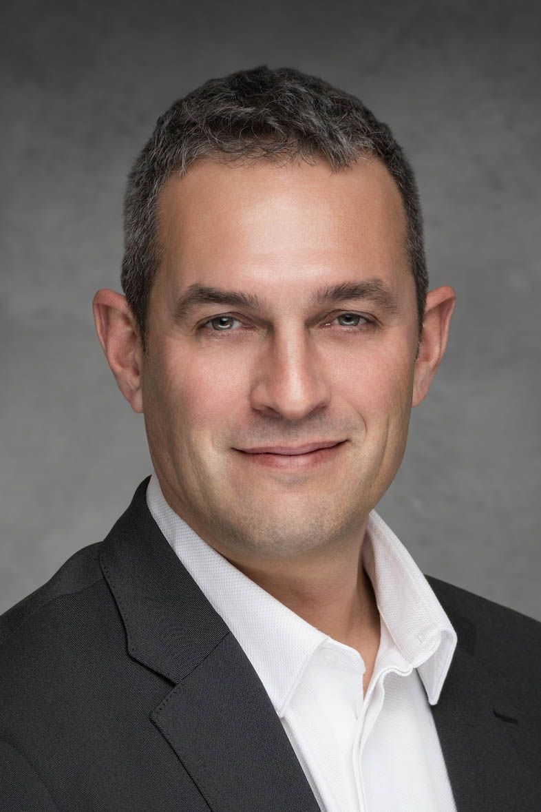 Professional headshot portrait of person in black suit jacket and white shirt against gray background.