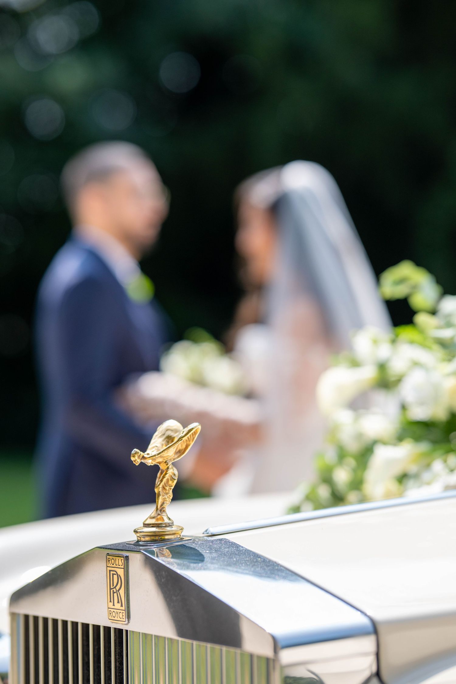 Close-up view of a Rolls-Royce Spirit of Ecstasy hood ornament with wedding couple blurred in background.