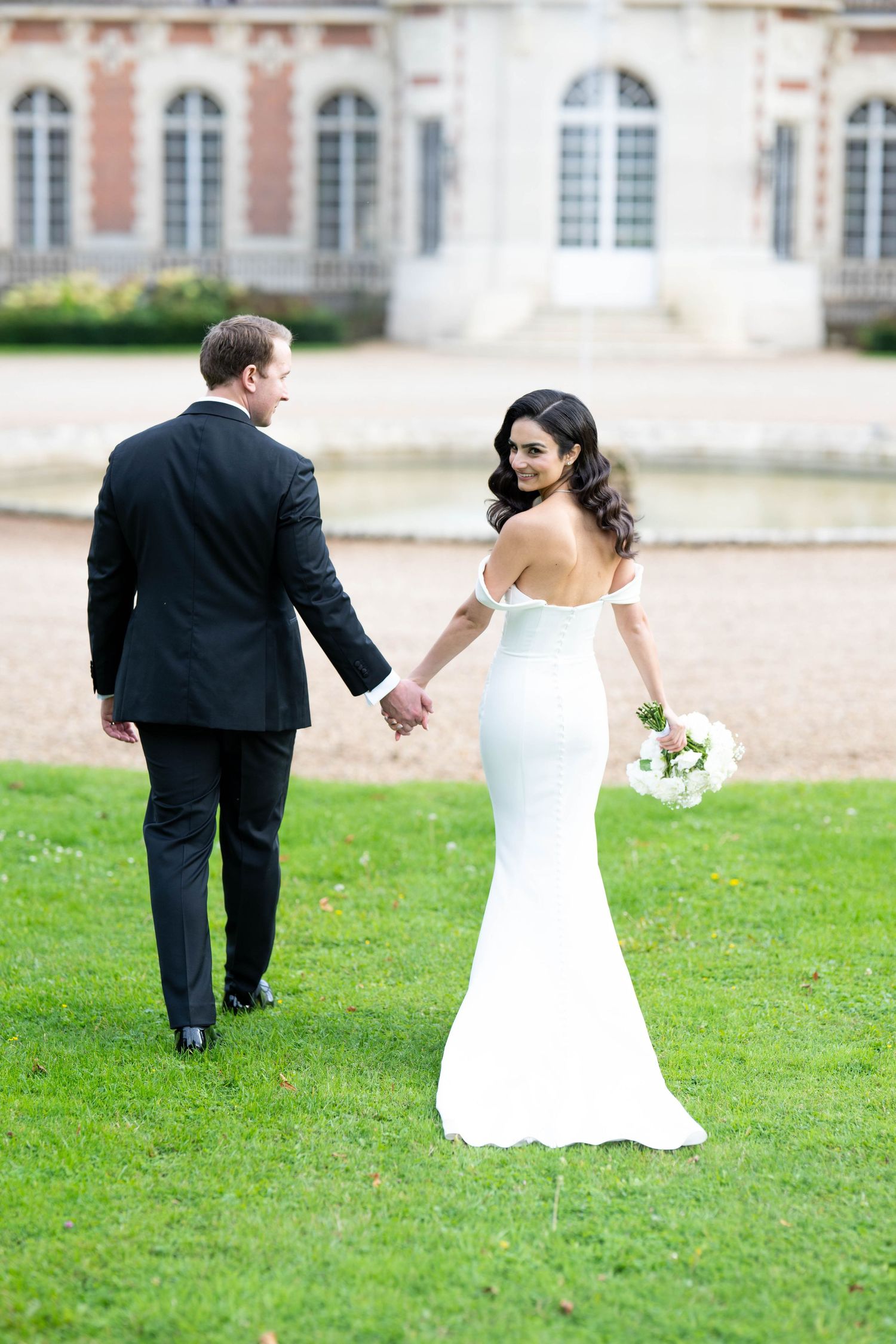 A bride and groom playfully walk across a green lawn in front of a historic building.