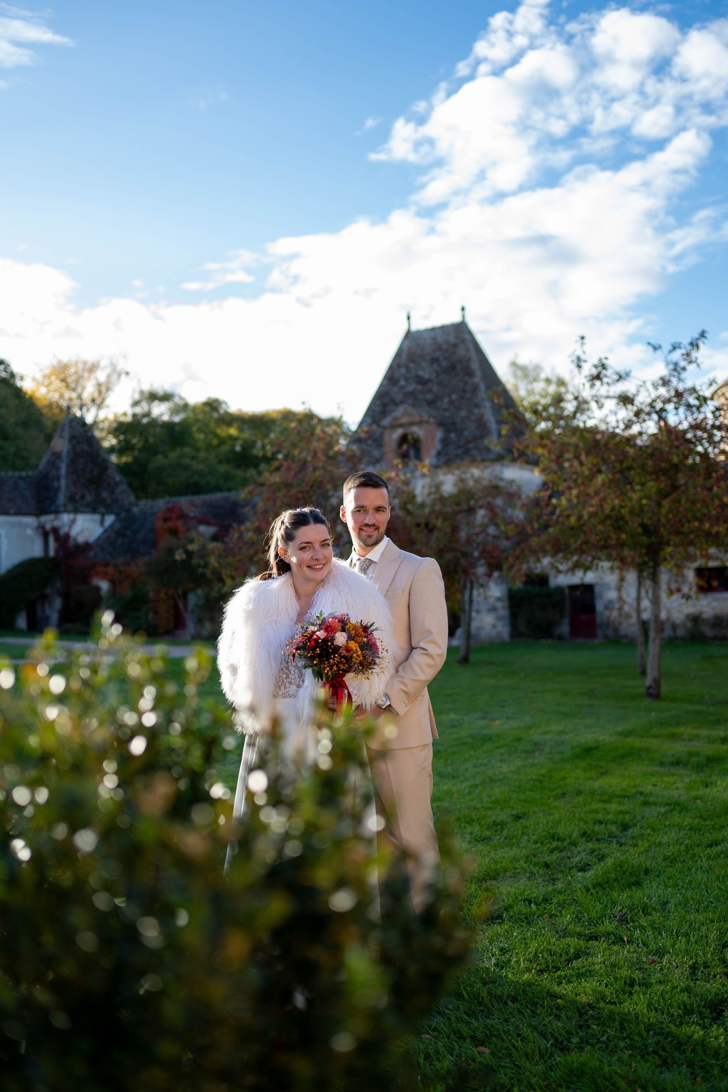 Wedding couple poses in a scenic outdoor garden setting with historic stone buildings in the background.