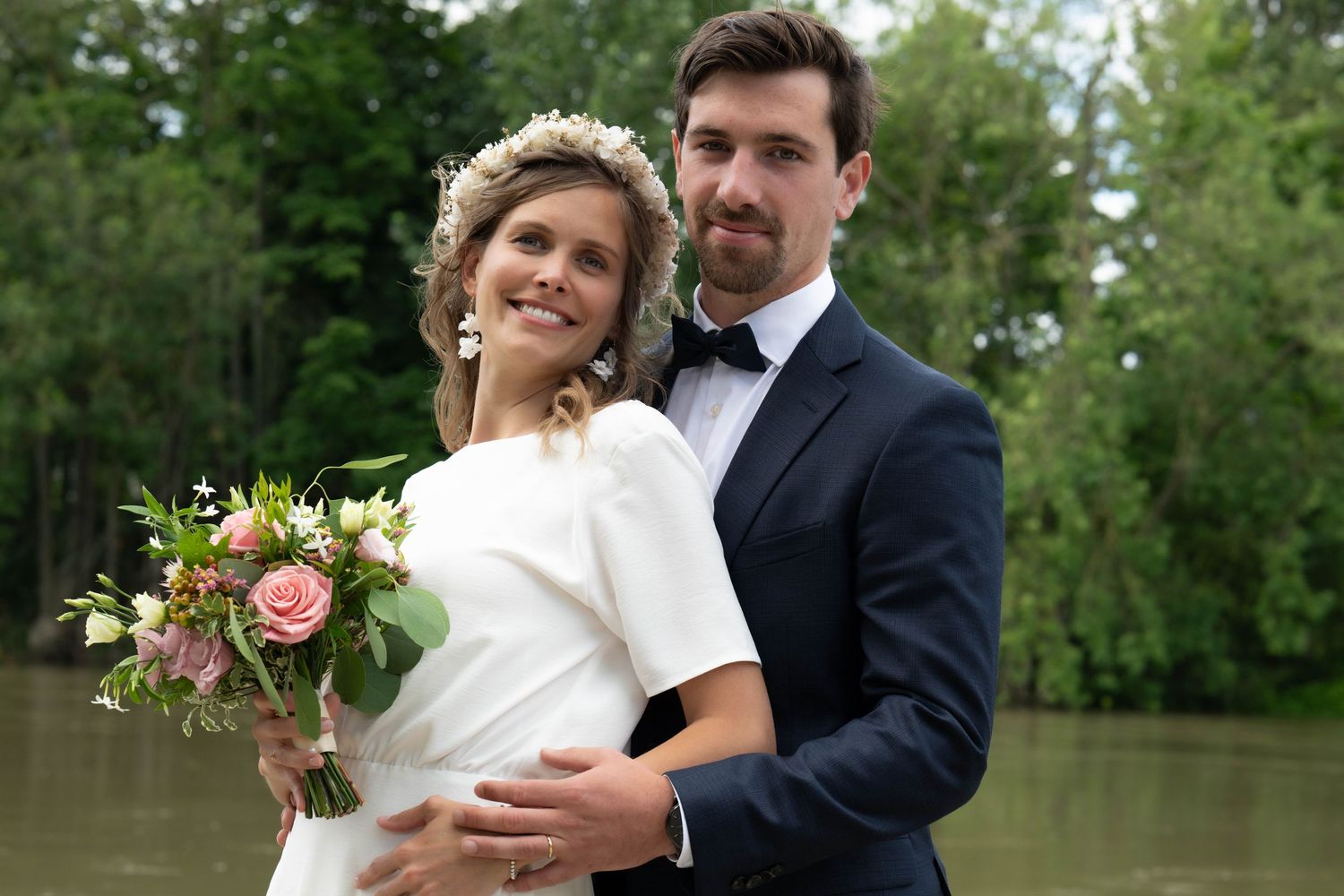 A romantic wedding couple embraces near a scenic lake surrounded by lush greenery during their outdoor celebration.