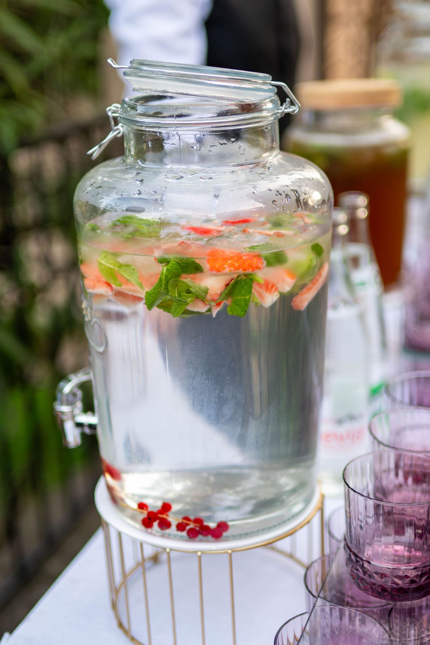 Large glass drink dispenser filled with infused water and garnished with fresh fruits and herbs.