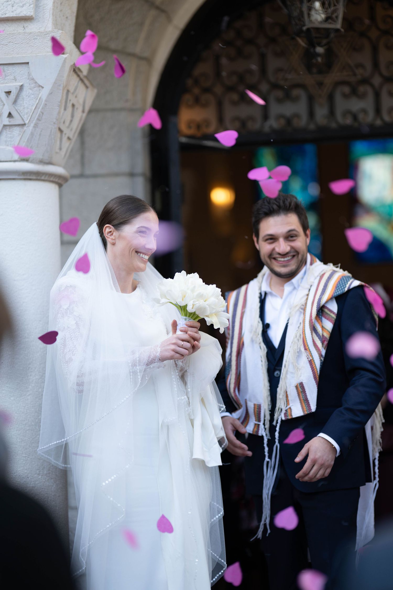 A joyful wedding couple shares a candid moment as pink flower petals fall around them in an outdoor setting.