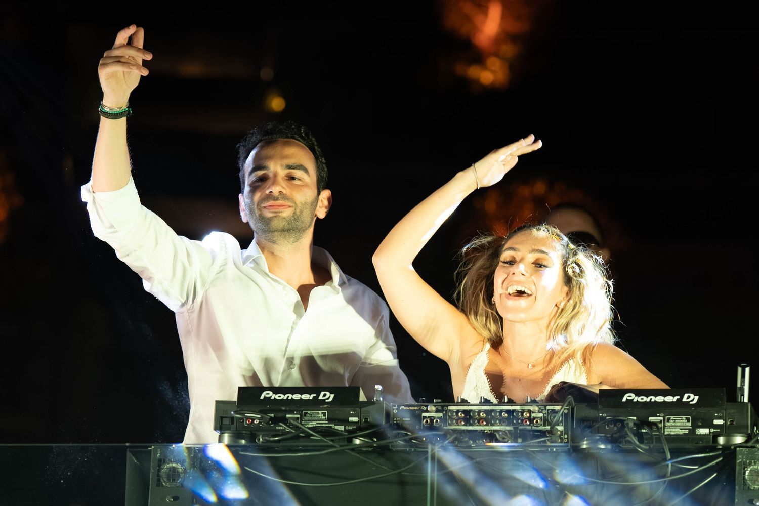 Two DJs perform energetically behind turntables at a nightclub with their arms raised in the air.