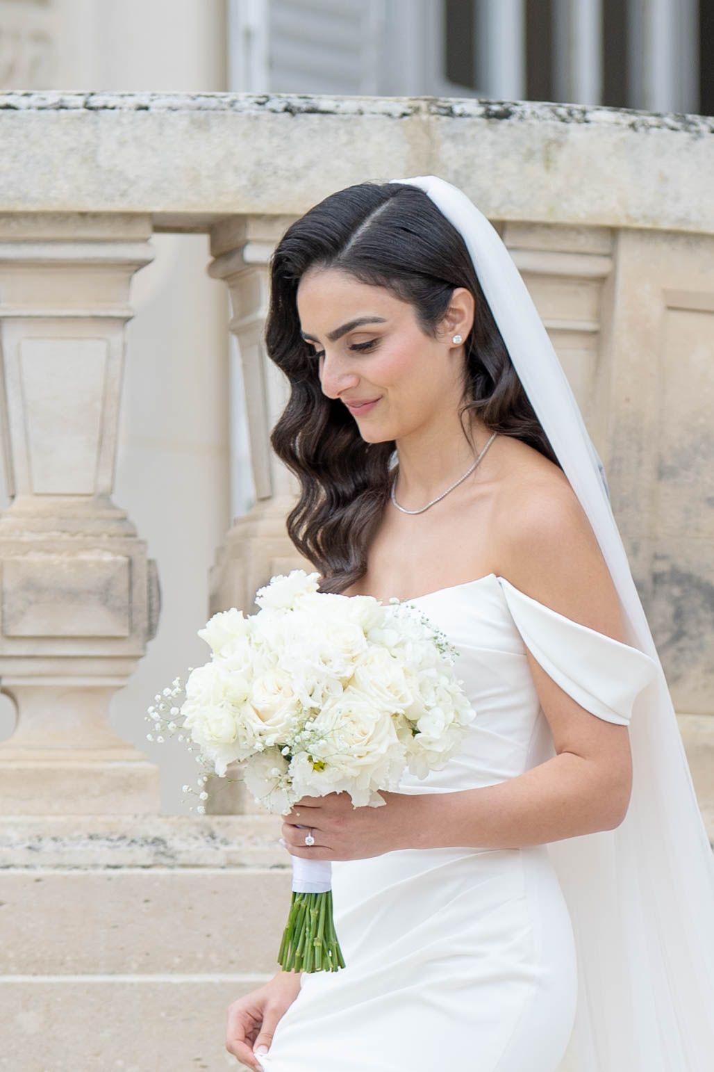 A bride in a white wedding dress holds a bouquet of white flowers against a light stone architectural background.