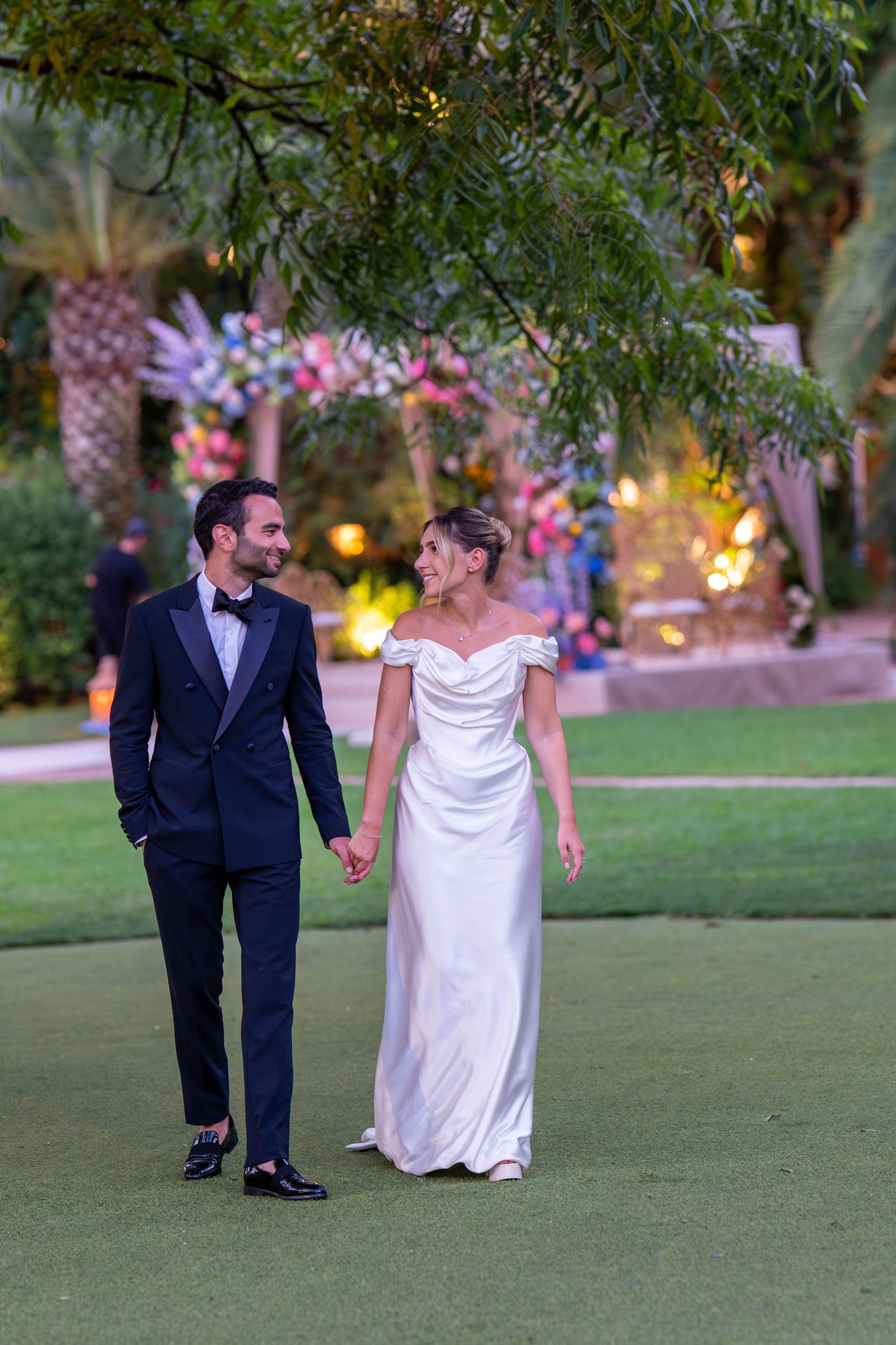 A couple walks hand in hand on a lawn with romantic lighting and floral decorations in the background.