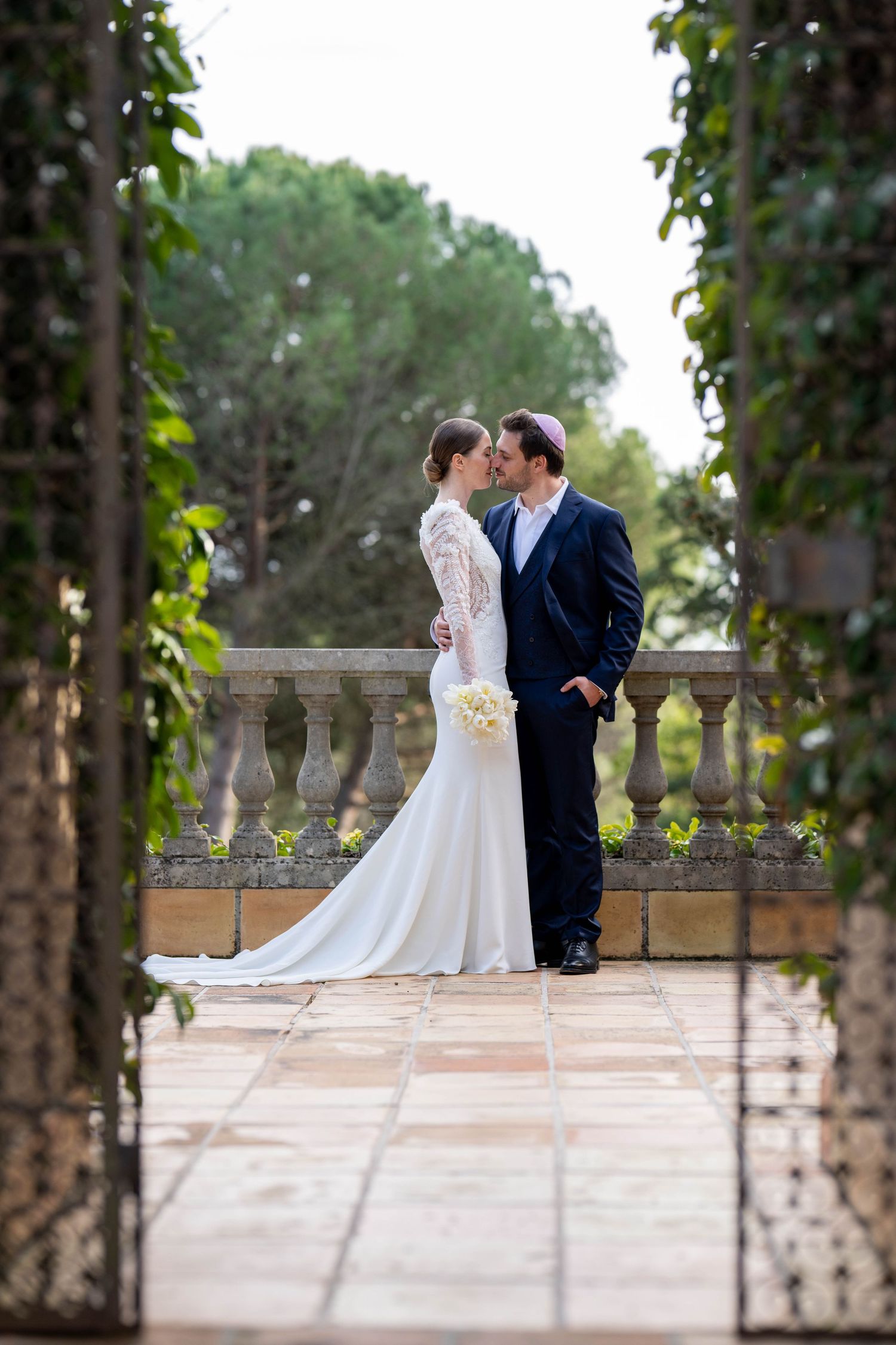 A romantic wedding couple embraces on an elegant stone terrace surrounded by lush greenery and ornate balustrades.