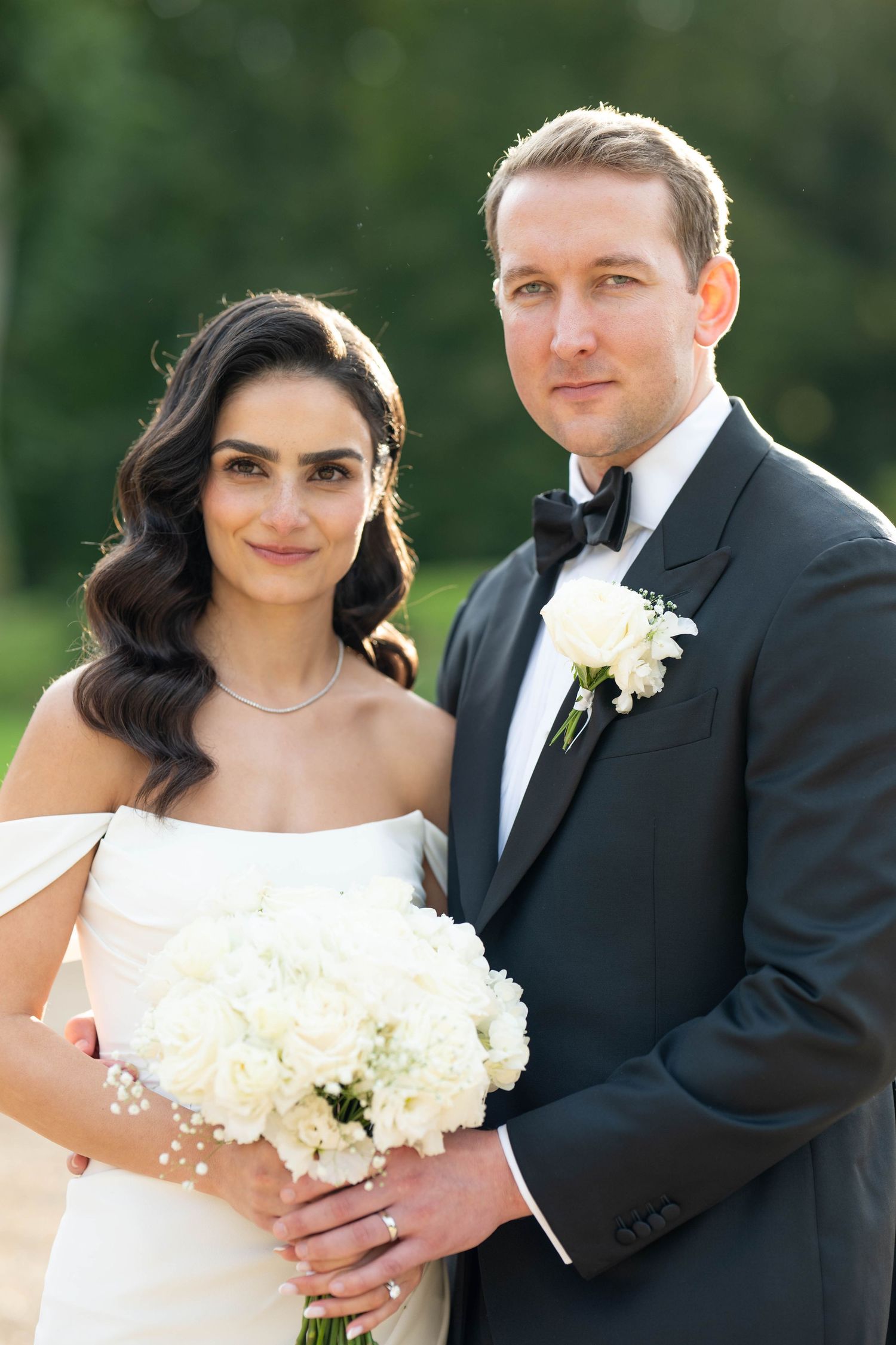 A bride and groom pose together outdoors in formal wedding attire with white floral bouquet.