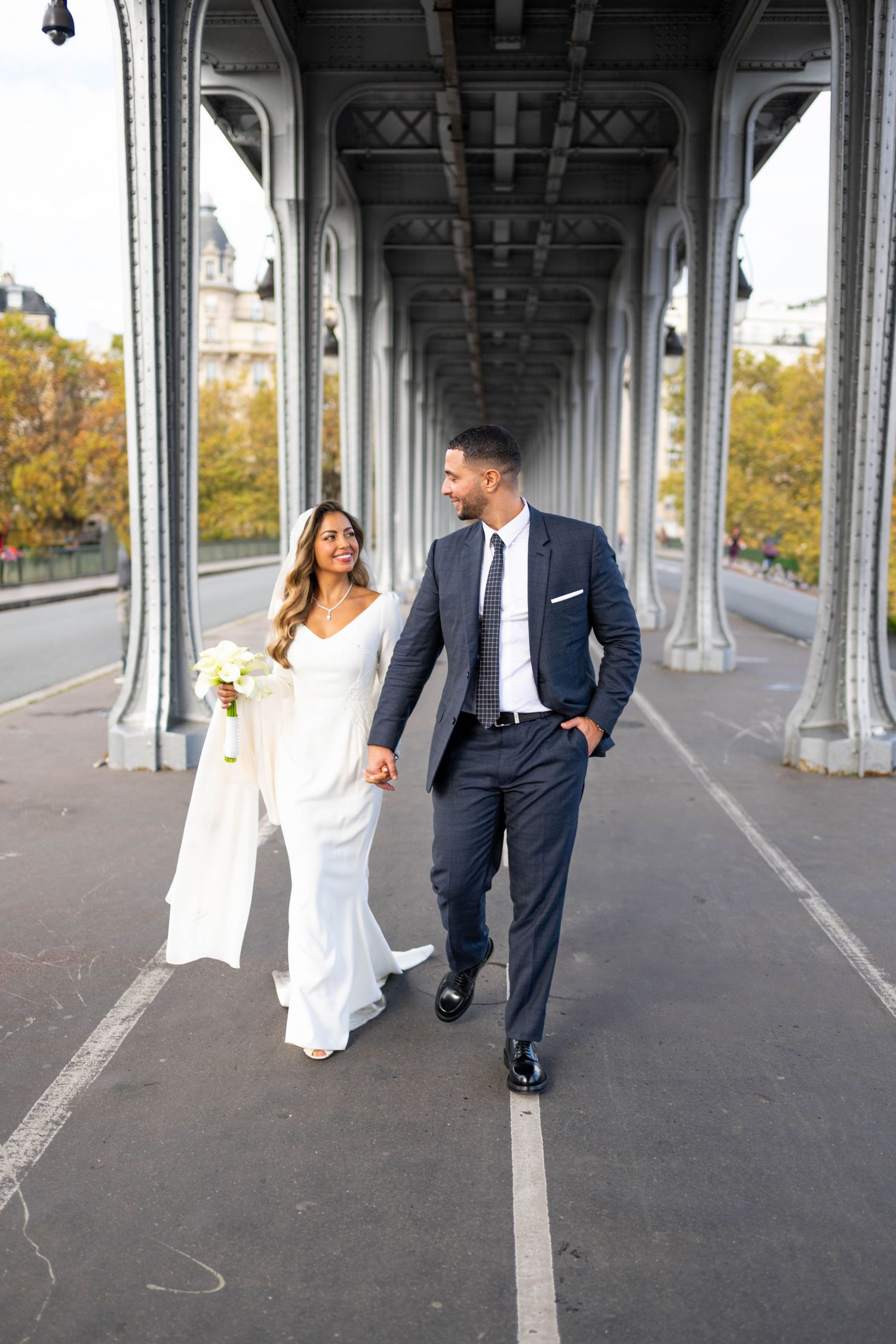 A couple in wedding attire walks and laughs together under a bridge archway during an autumn day.