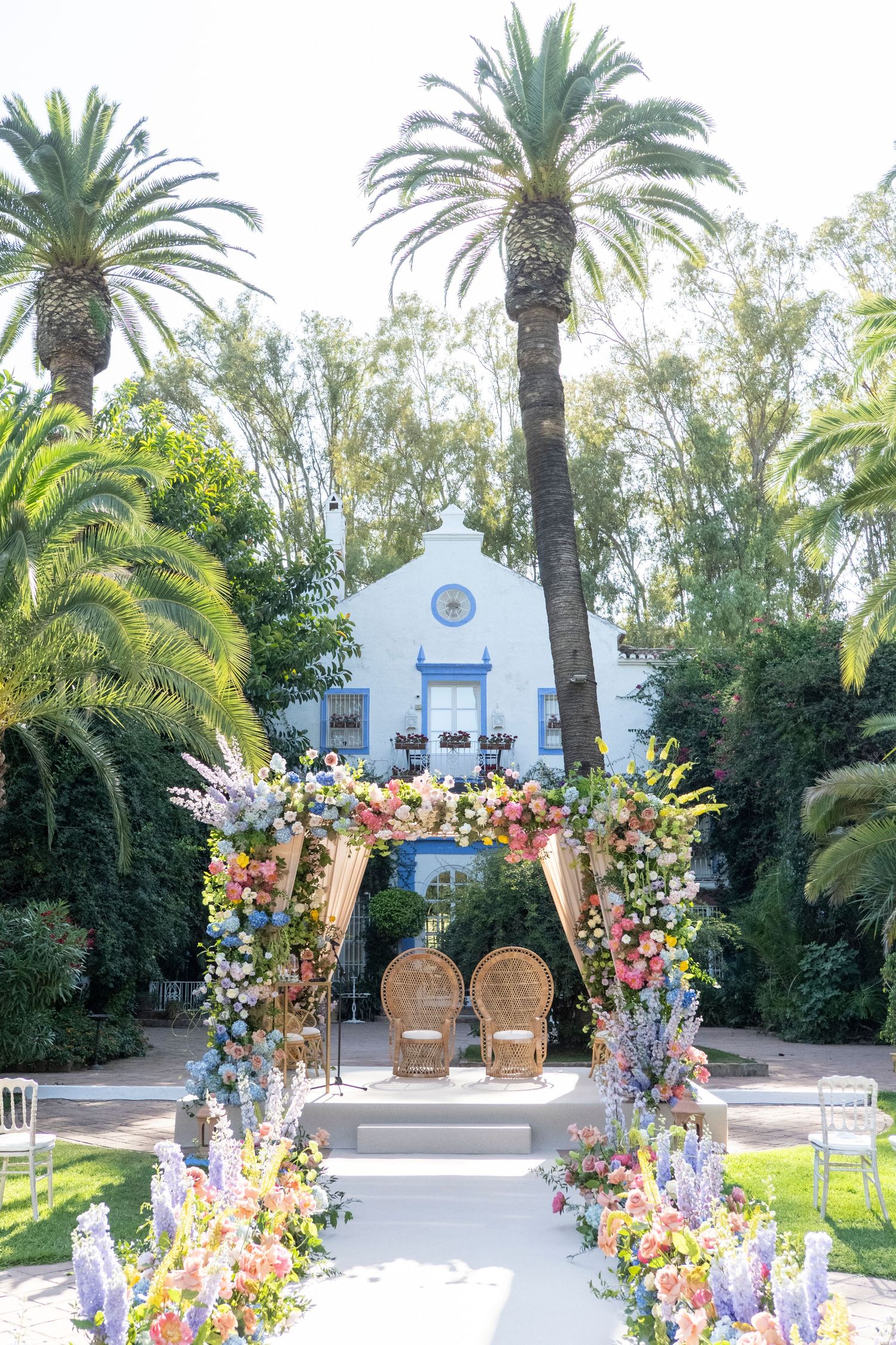 Outdoor wedding ceremony setup with colorful floral arch and palm trees at white chapel.