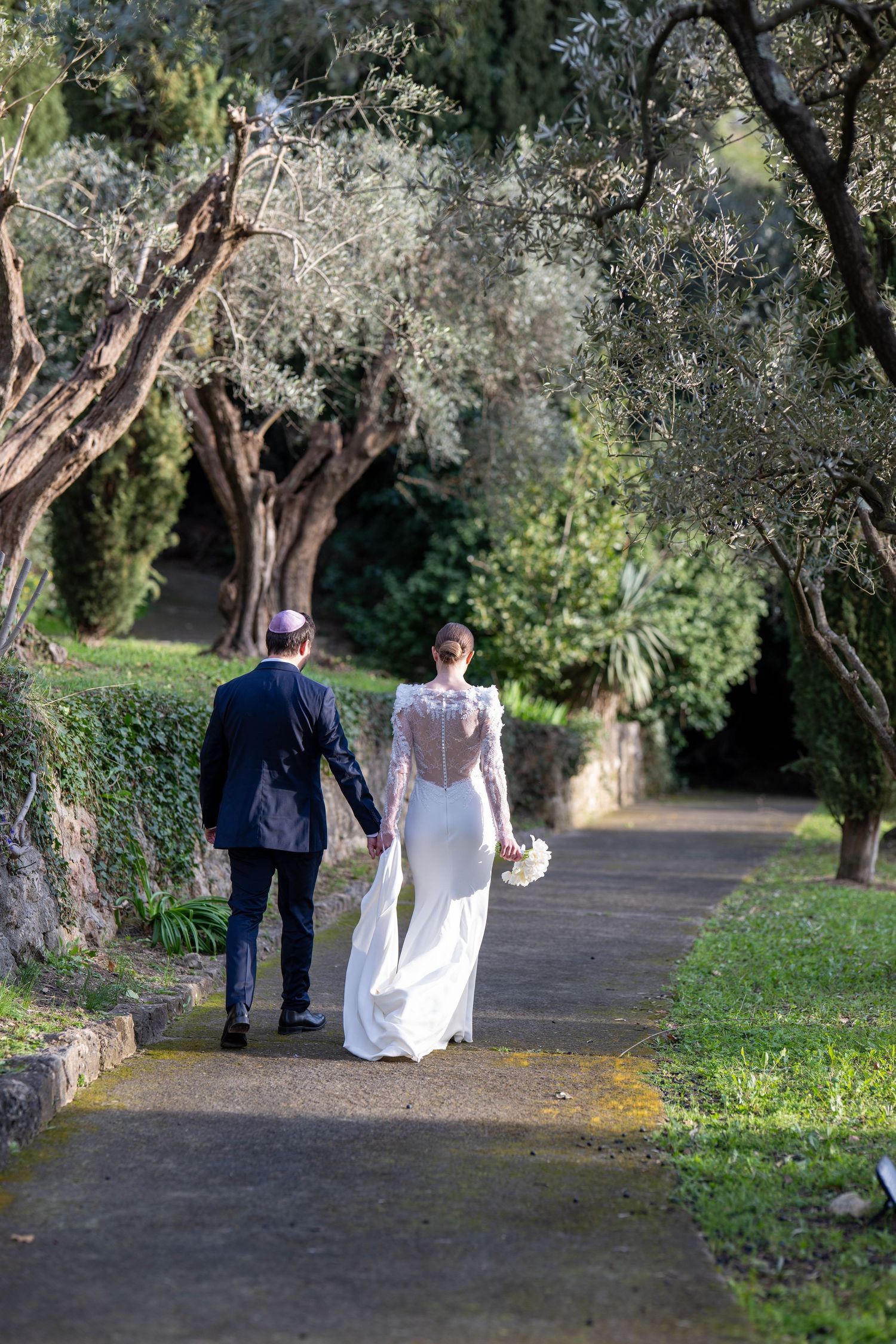 Couple walking through olive grove garden path after wedding ceremony.