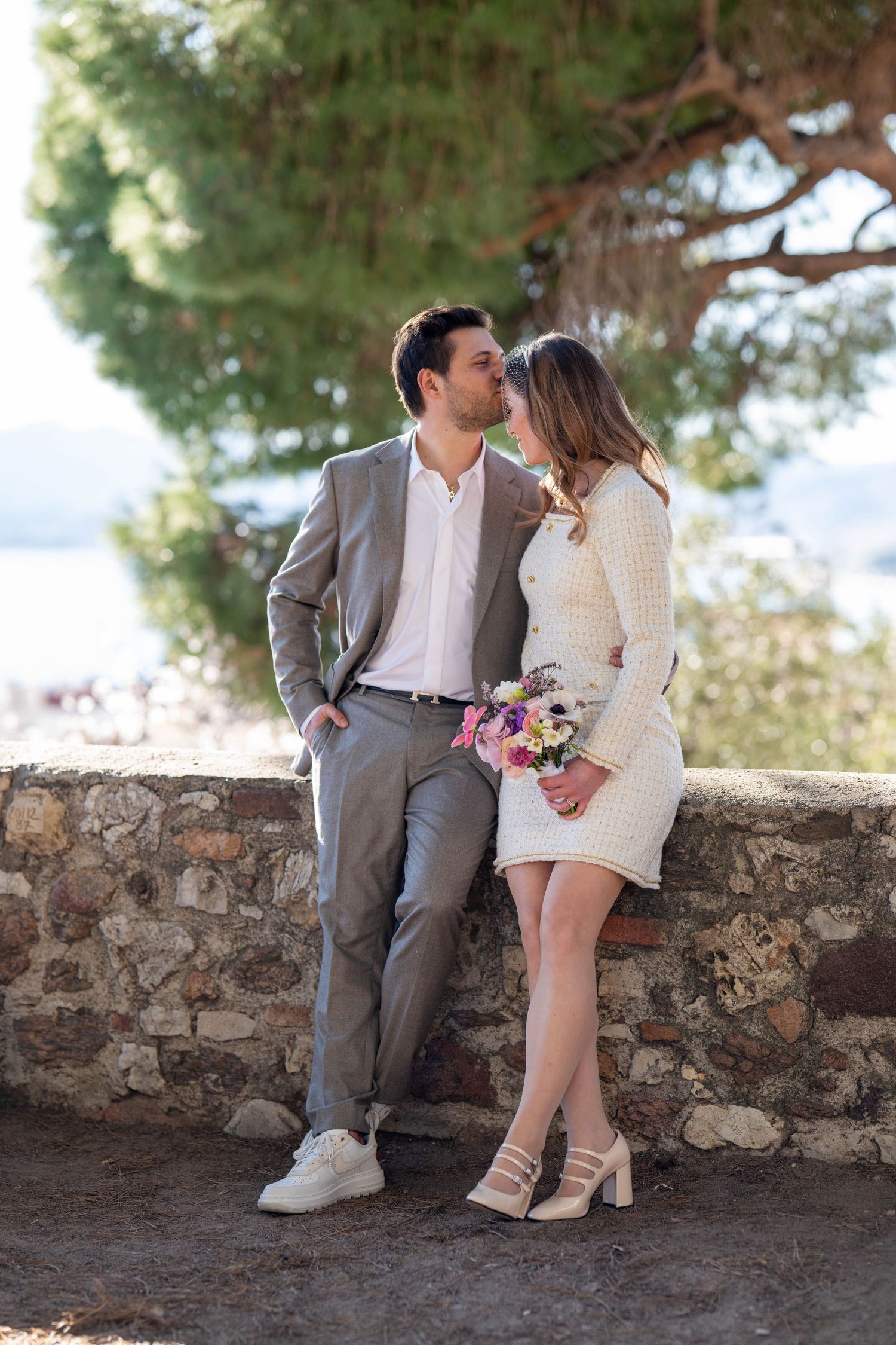 A romantic couple sits on a stone wall sharing intimate moments in a Mediterranean setting with pine trees and sea views.