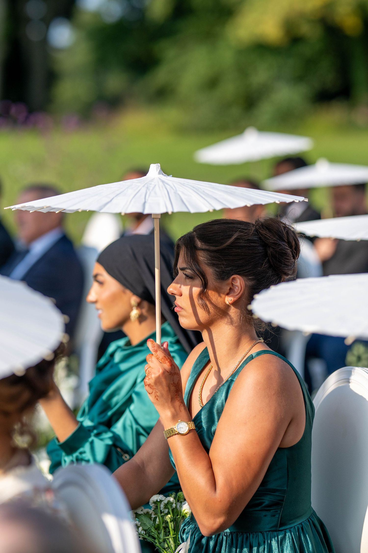 Row of guests in teal dresses holding white parasols at an outdoor wedding ceremony.