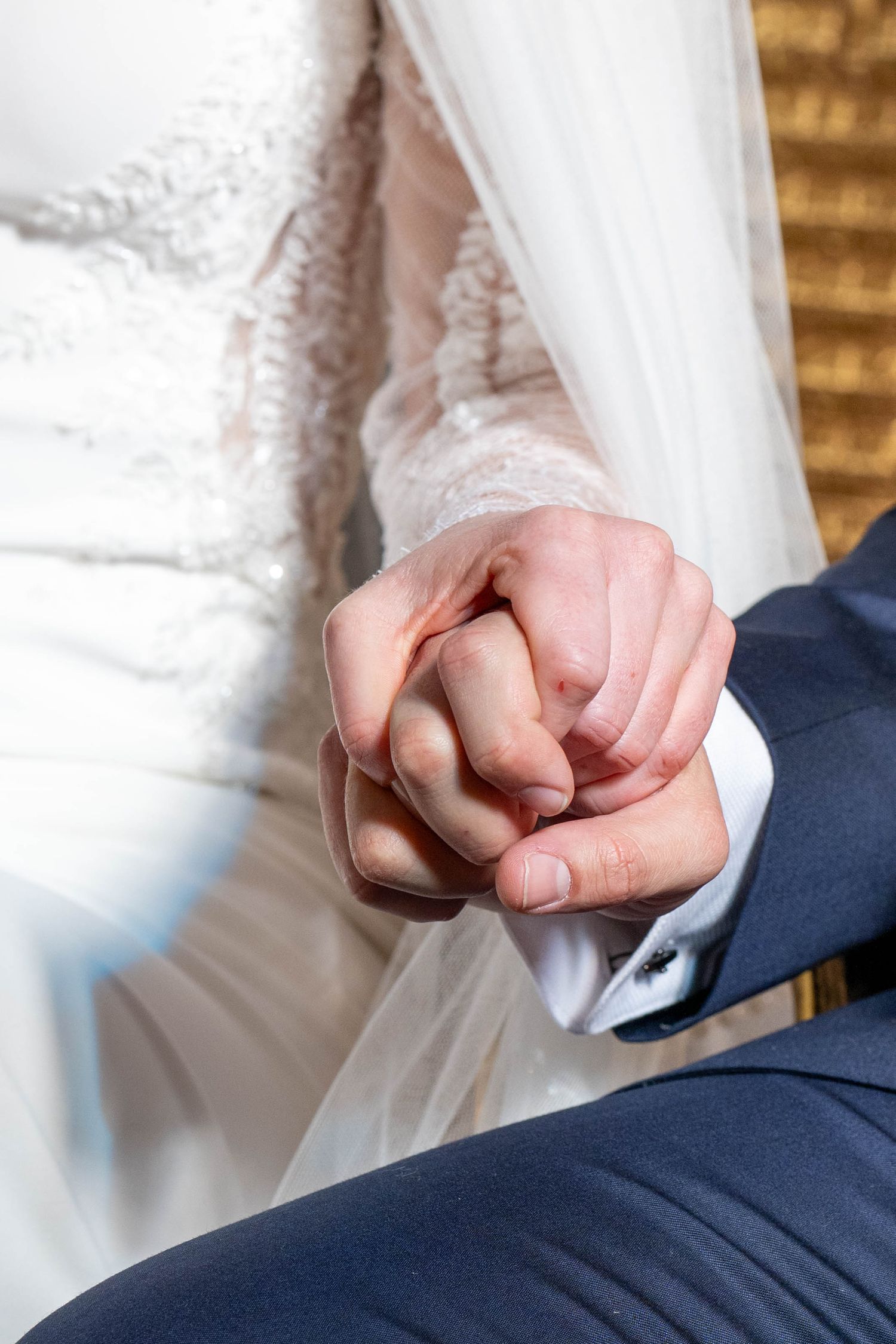 Close-up of a couple holding hands at a wedding, showing white lace dress and navy suit details.