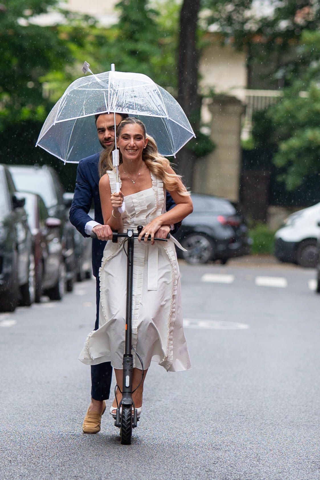 A couple shares a clear umbrella while walking down a rainy street in casual attire.